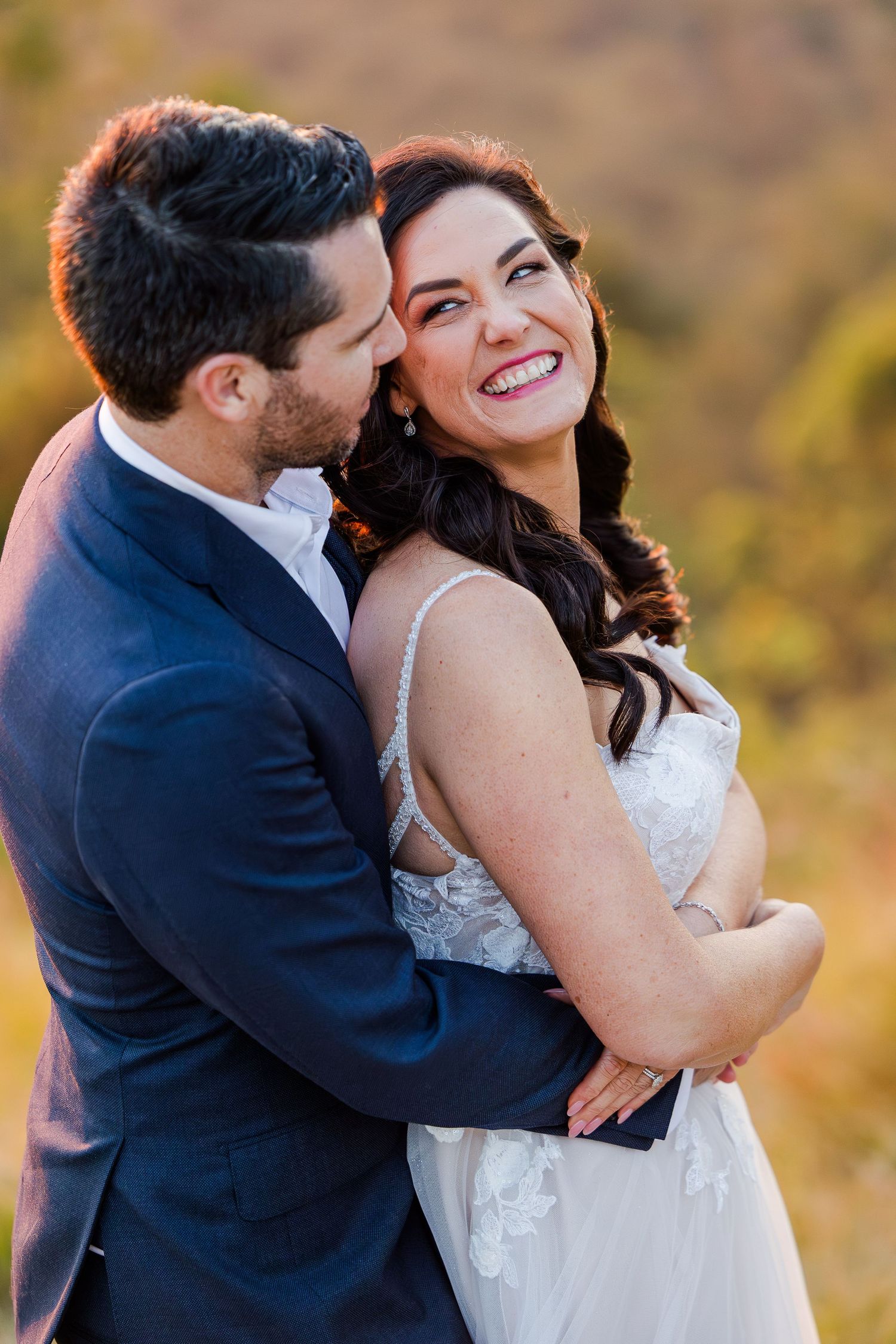 A romantic outdoor wedding portrait showing a couple embracing and smiling in warm autumn light.