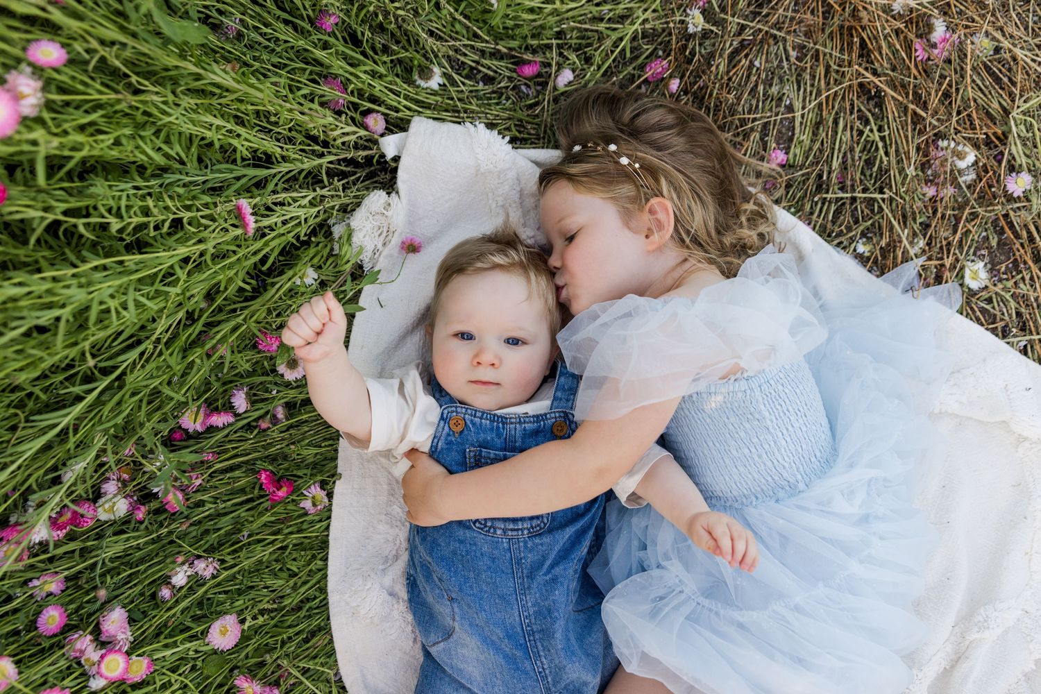 Baby in denim dress and white bow lies on white blanket surrounded by greenery and pink flower petals.