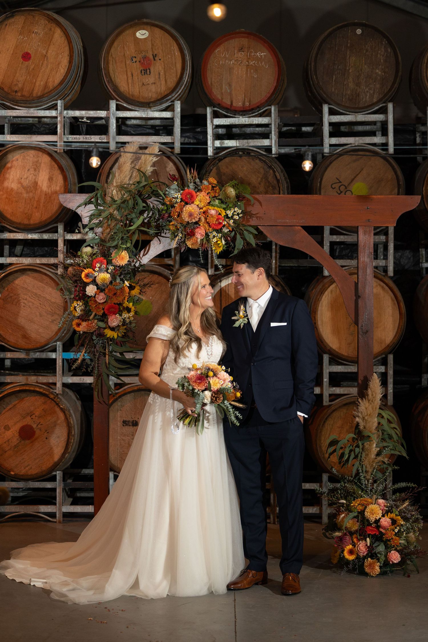 Bride and groom pose before wine barrel wall with vibrant fall floral arch in warm oranges and yellows at winery wedding.