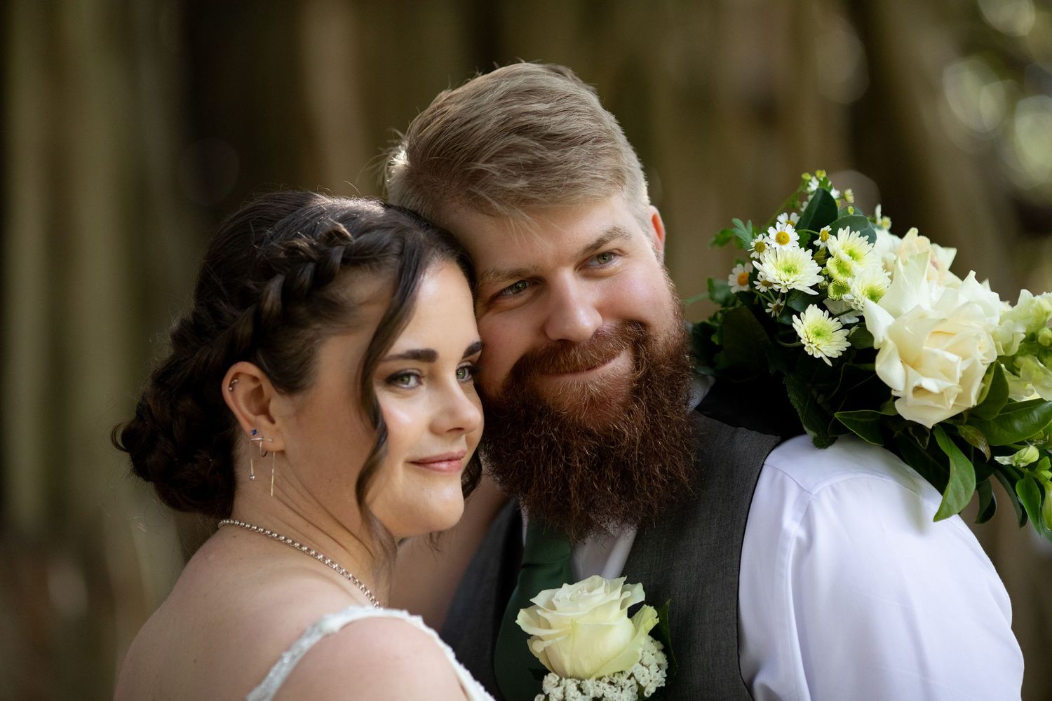 Smiling bride and bearded groom pose together holding white floral bouquet on their wedding day.