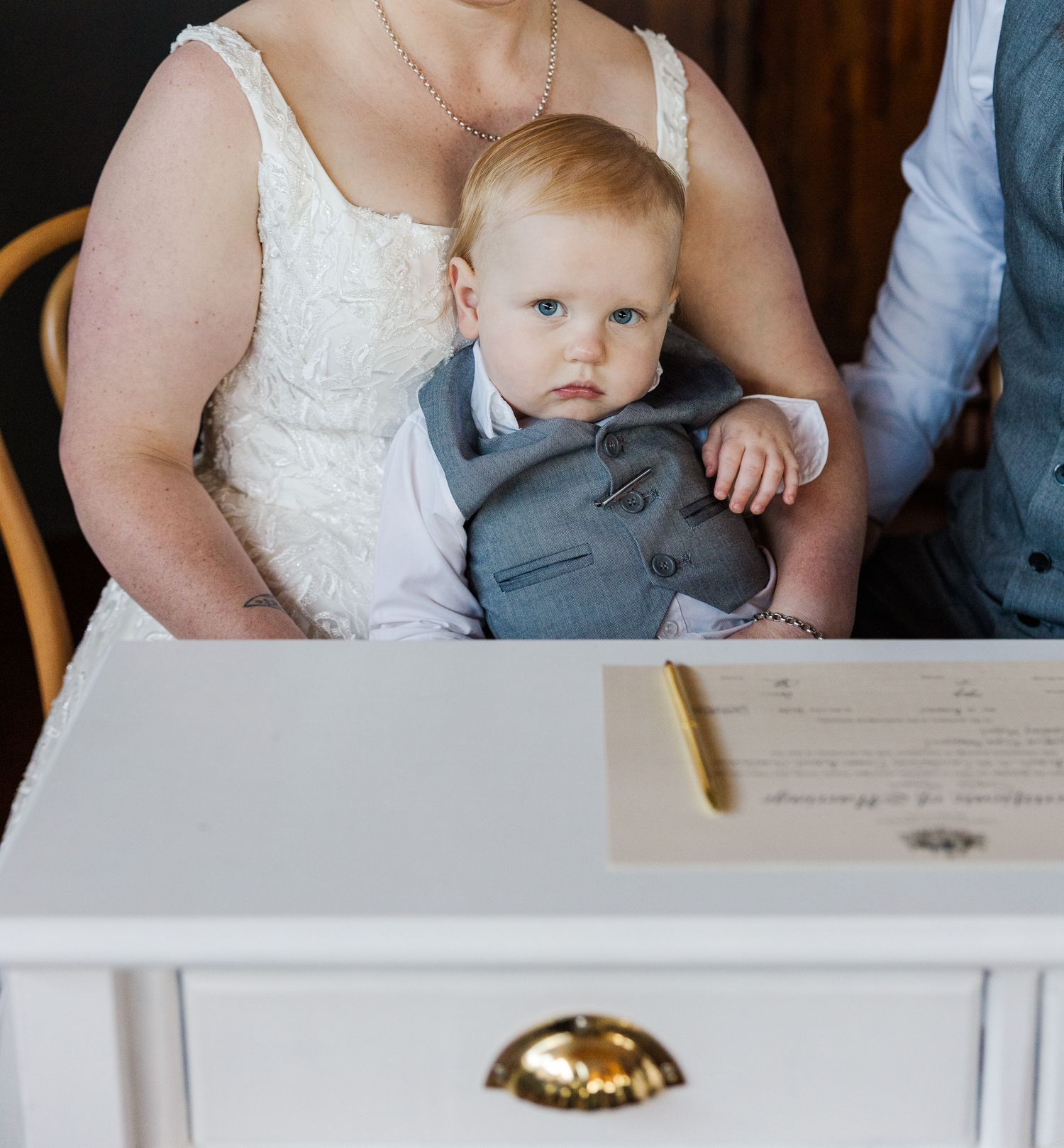 Baby in gray suit held by bride in white lace dress at wedding ceremony signing table.