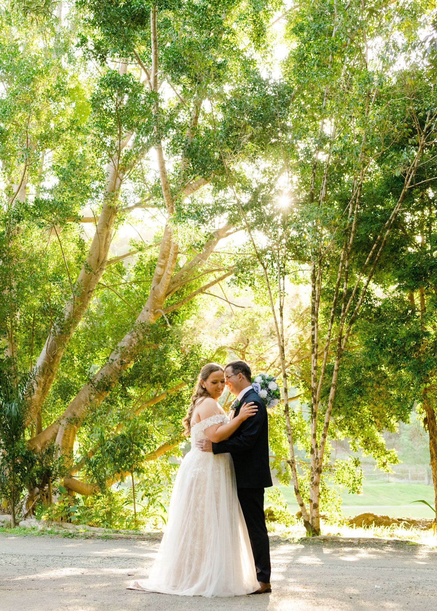 A romantic moment captured under sunlit trees in a serene outdoor setting with a white wedding dress and formal attire.