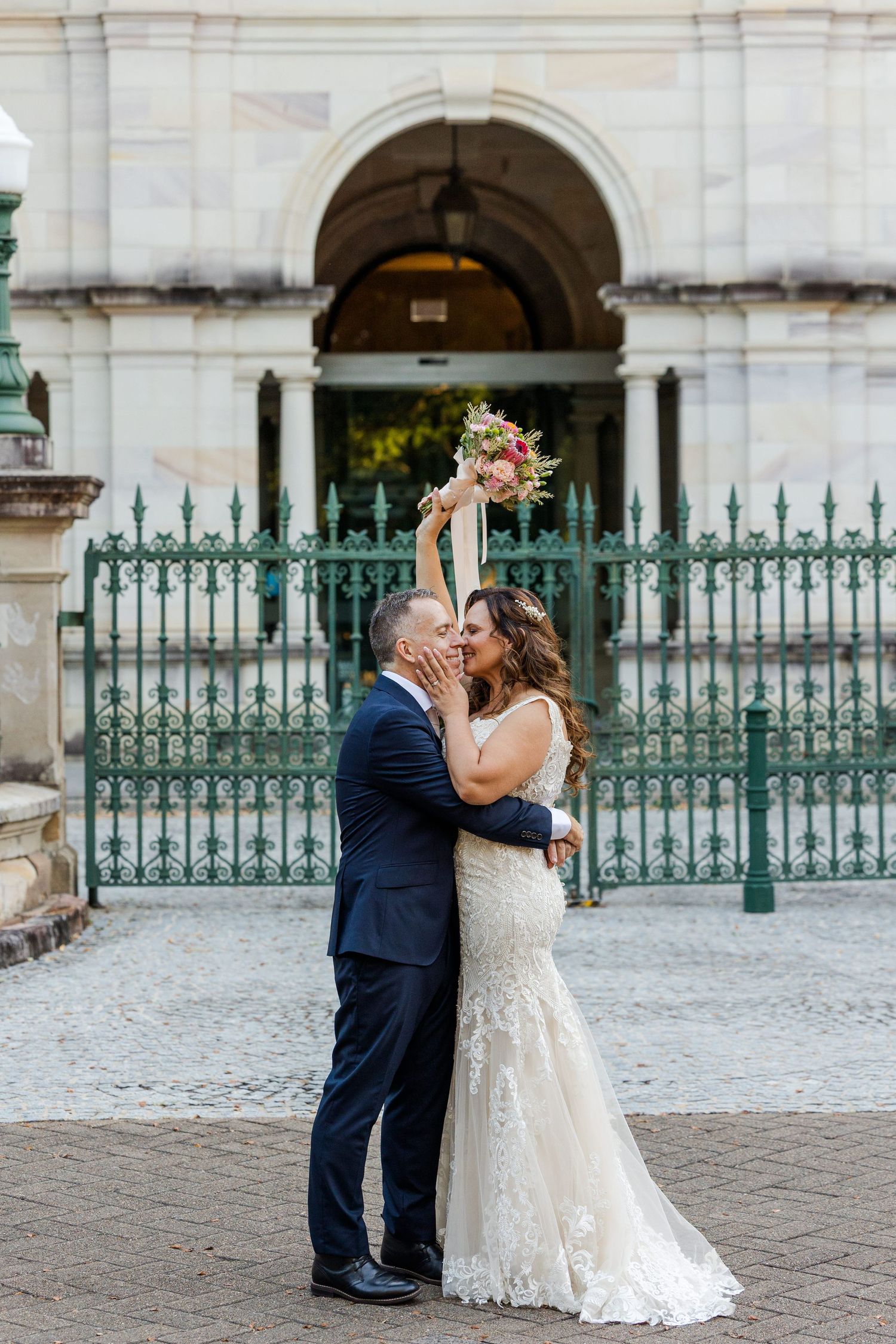 A couple embraces on cobblestone pavement in front of an ornate iron gate and stone archway.