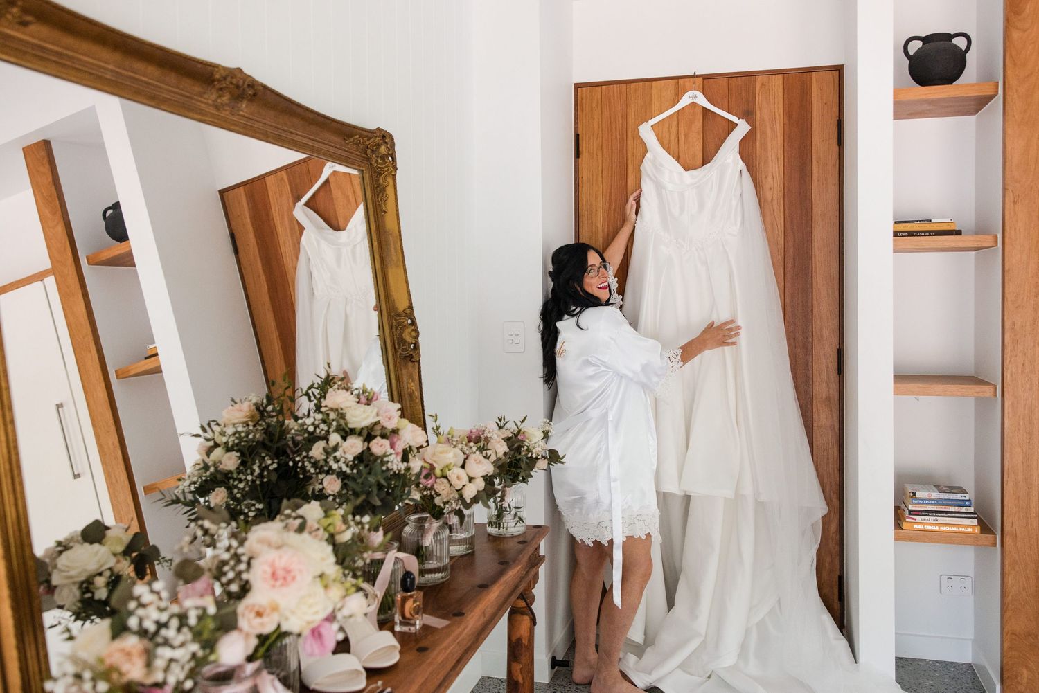 Bride in white robe admiring her wedding gown hanging on display, with floral arrangements and a gold mirror nearby.