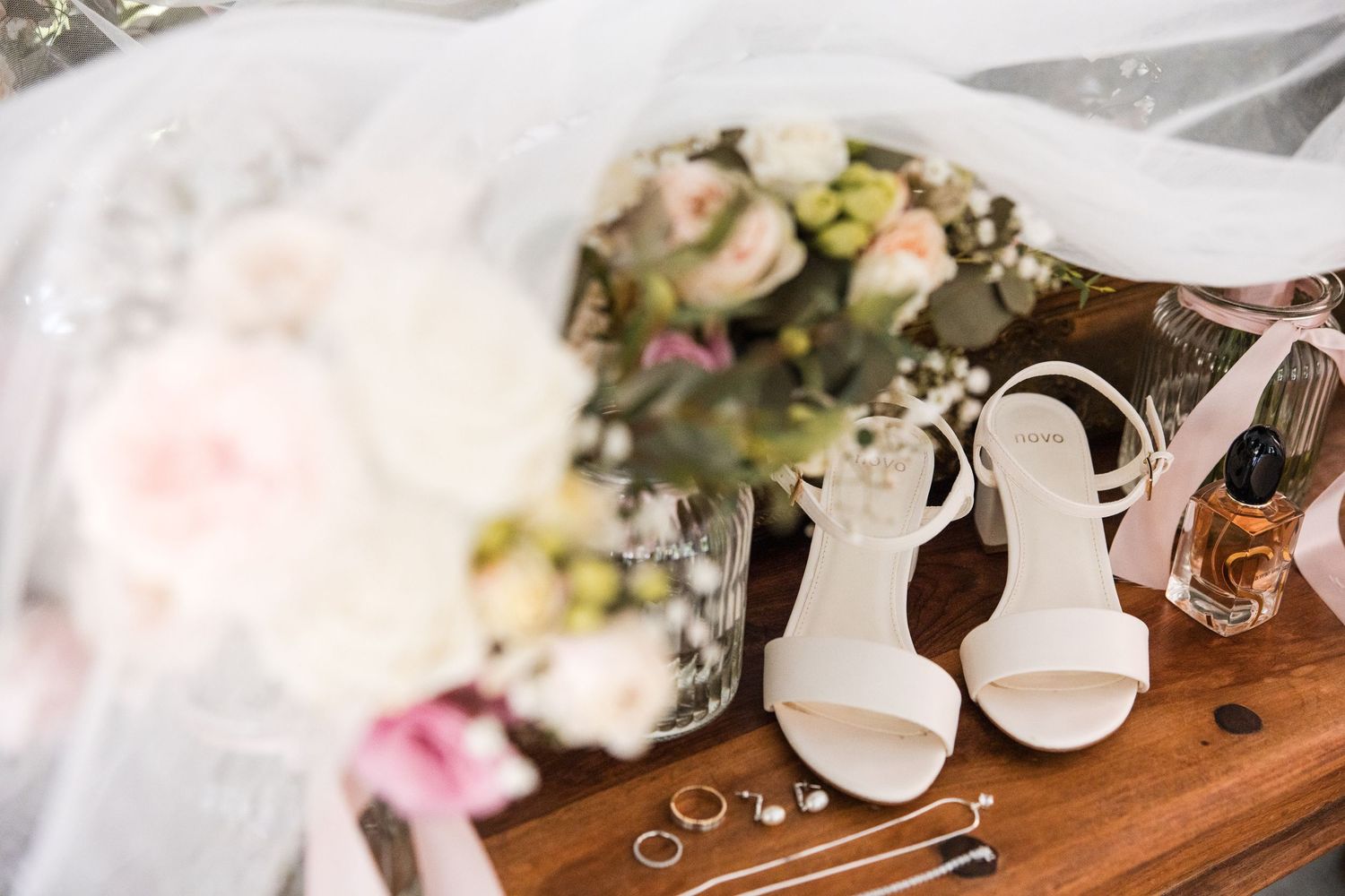 Elegant wedding flat lay with white bridal heels, floral bouquet, gold rings, and perfume on a wooden surface.