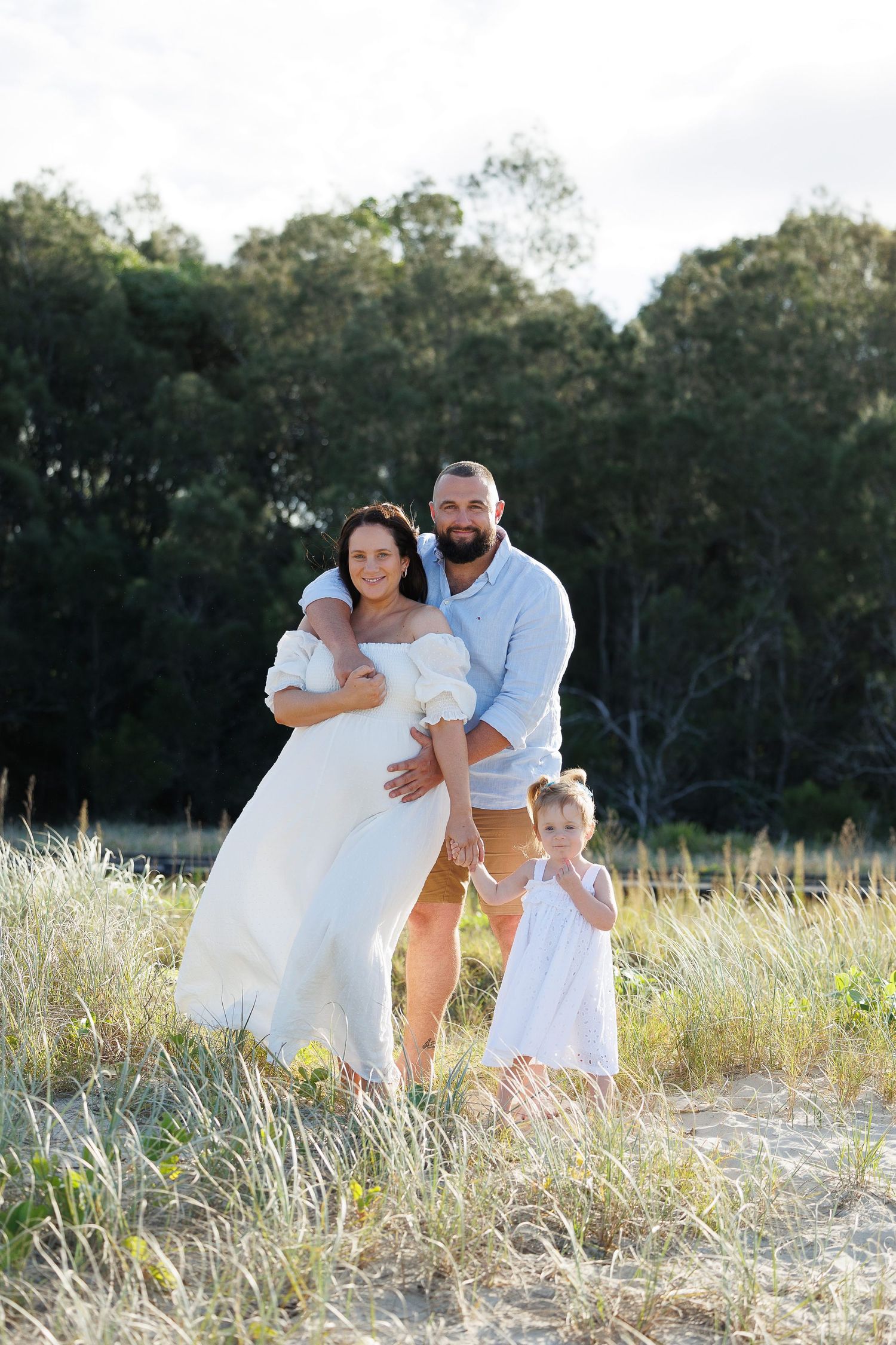 A family wearing coordinated white outfits poses together in a sunny field with trees in the background.
