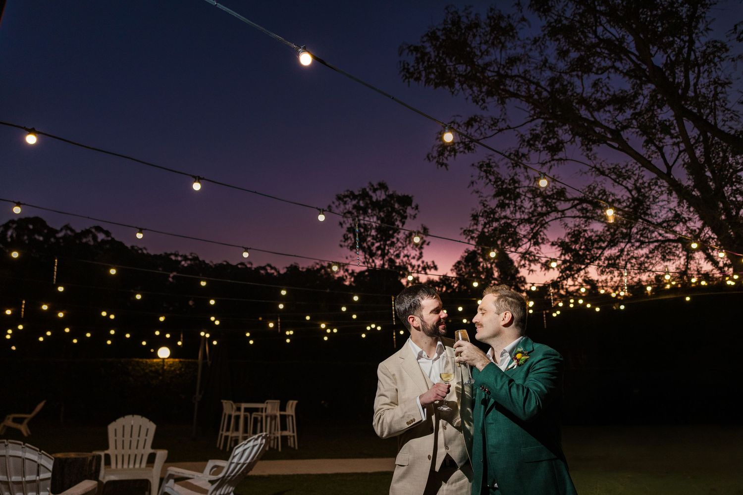 Two grooms share a romantic moment under string lights at twilight during an outdoor wedding reception.