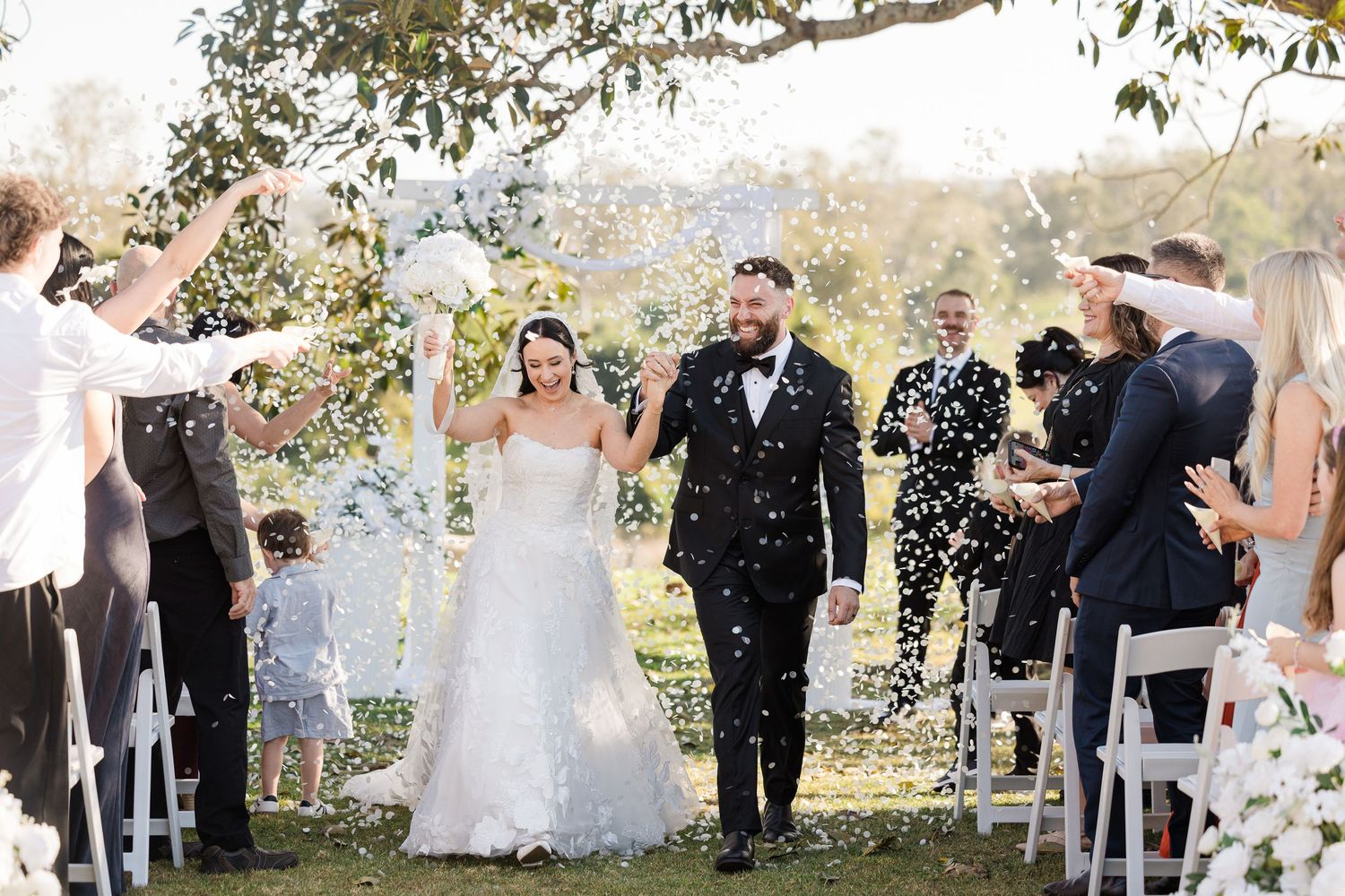 Bride and groom walk down outdoor aisle as guests toss white confetti at sunny wedding ceremony.