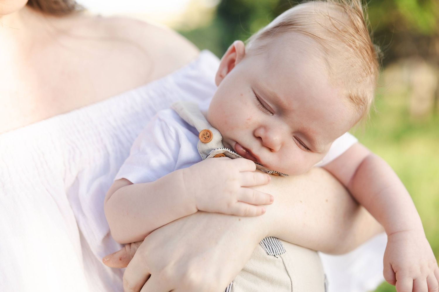Peaceful sleeping baby resting on mother's shoulder outdoors in warm natural light, wearing a white onesie.