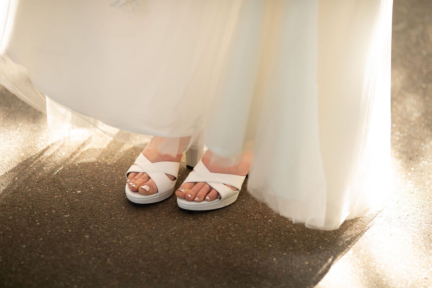 Close-up of a bride wearing white cross-strap sandals beneath a flowing white wedding gown on a sunny day.