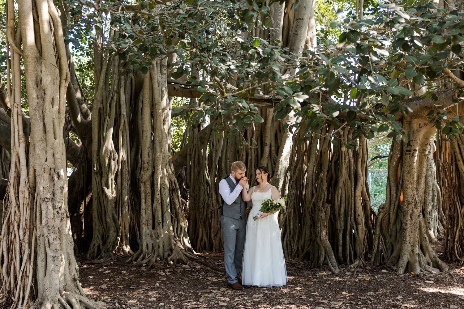 Bride and groom standing beneath a massive banyan tree with aerial roots during an outdoor wedding ceremony.