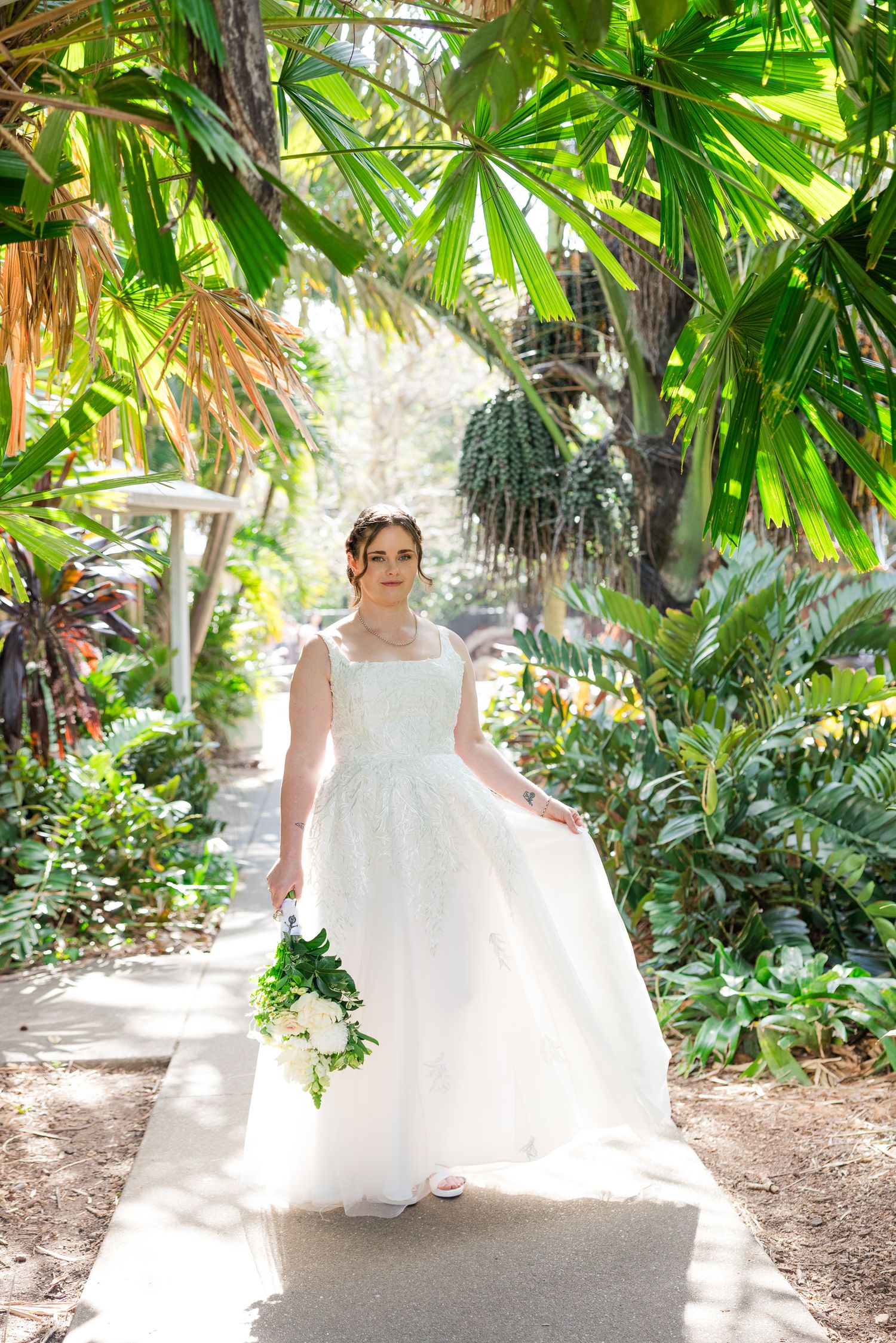 Bride in white gown holds bouquet on garden path surrounded by lush tropical palms and greenery.