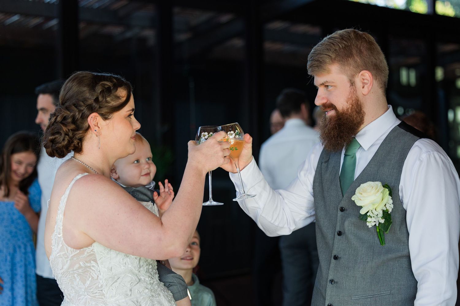 Bride holding baby toasts with groom in gray vest and green tie at outdoor wedding reception.