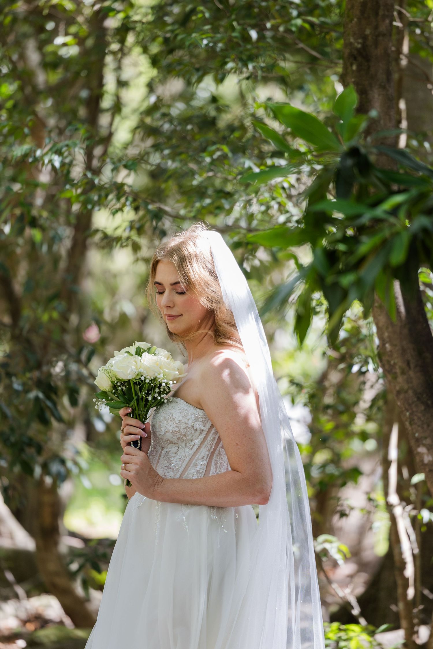 A bride in a white strapless gown holds her bouquet in a sunlit garden setting.