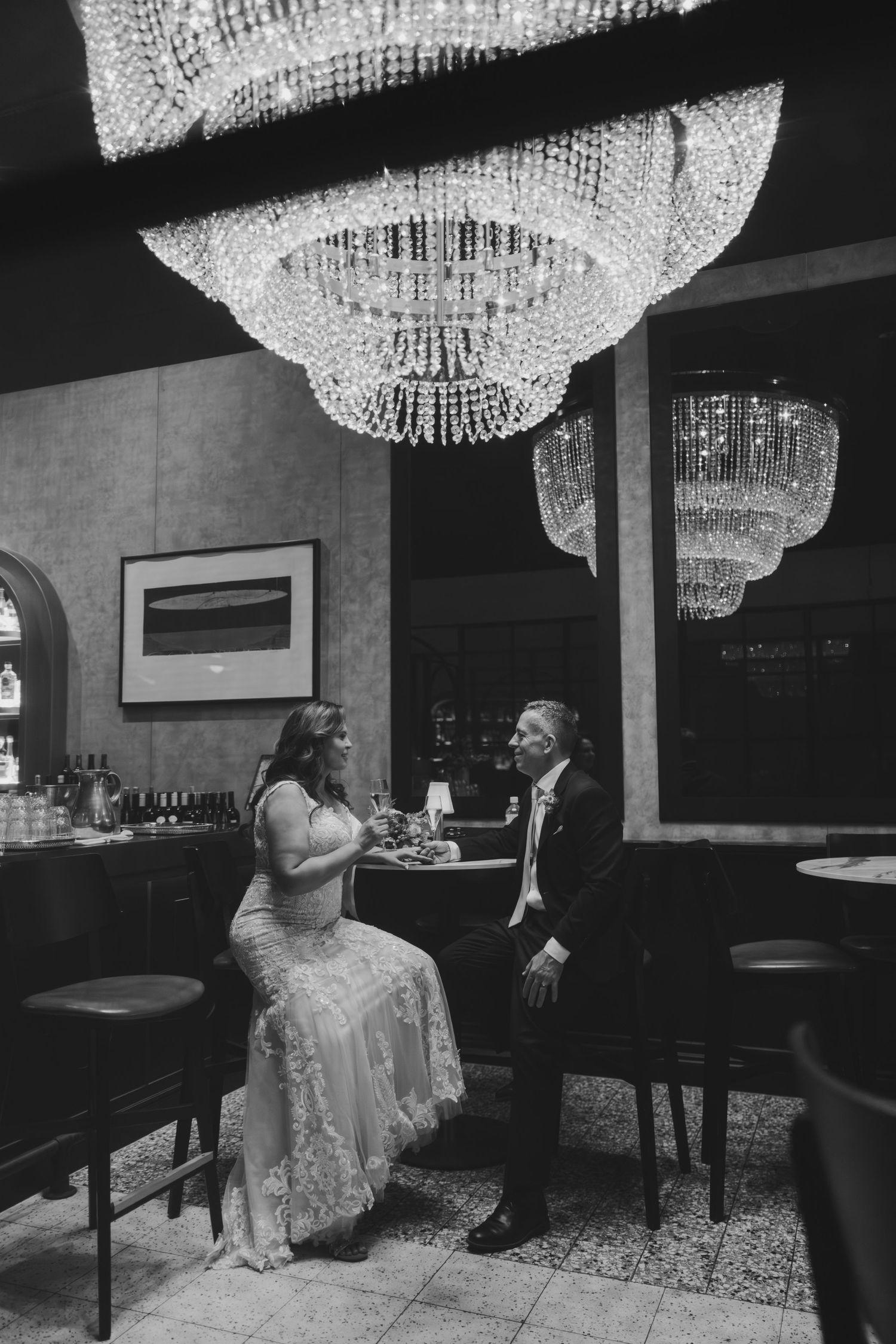 A couple shares an intimate moment at an elegant bar beneath ornate crystal chandeliers in black and white photography.