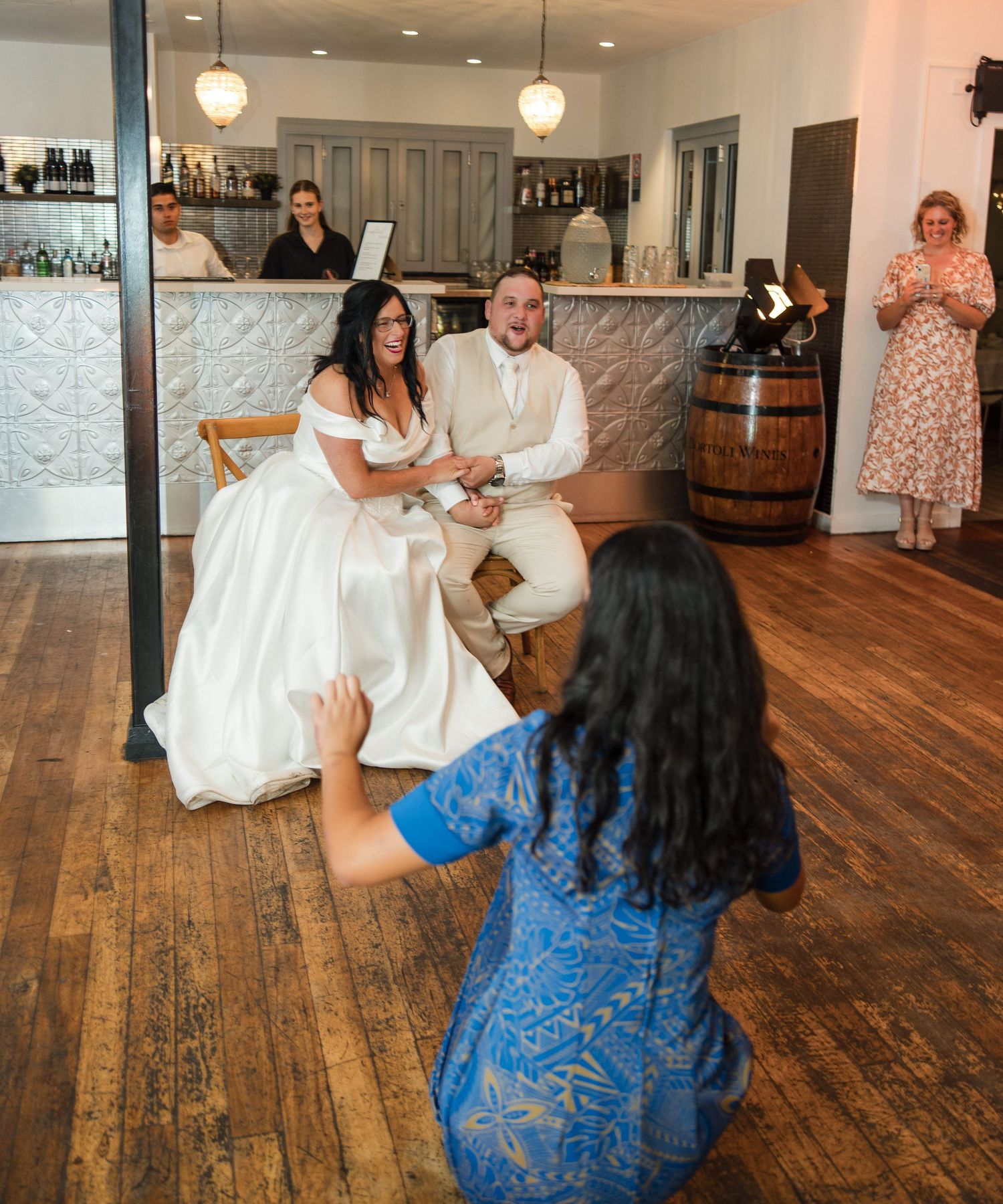 Bride and groom seated at wedding reception as guest in blue dress approaches them on dance floor.