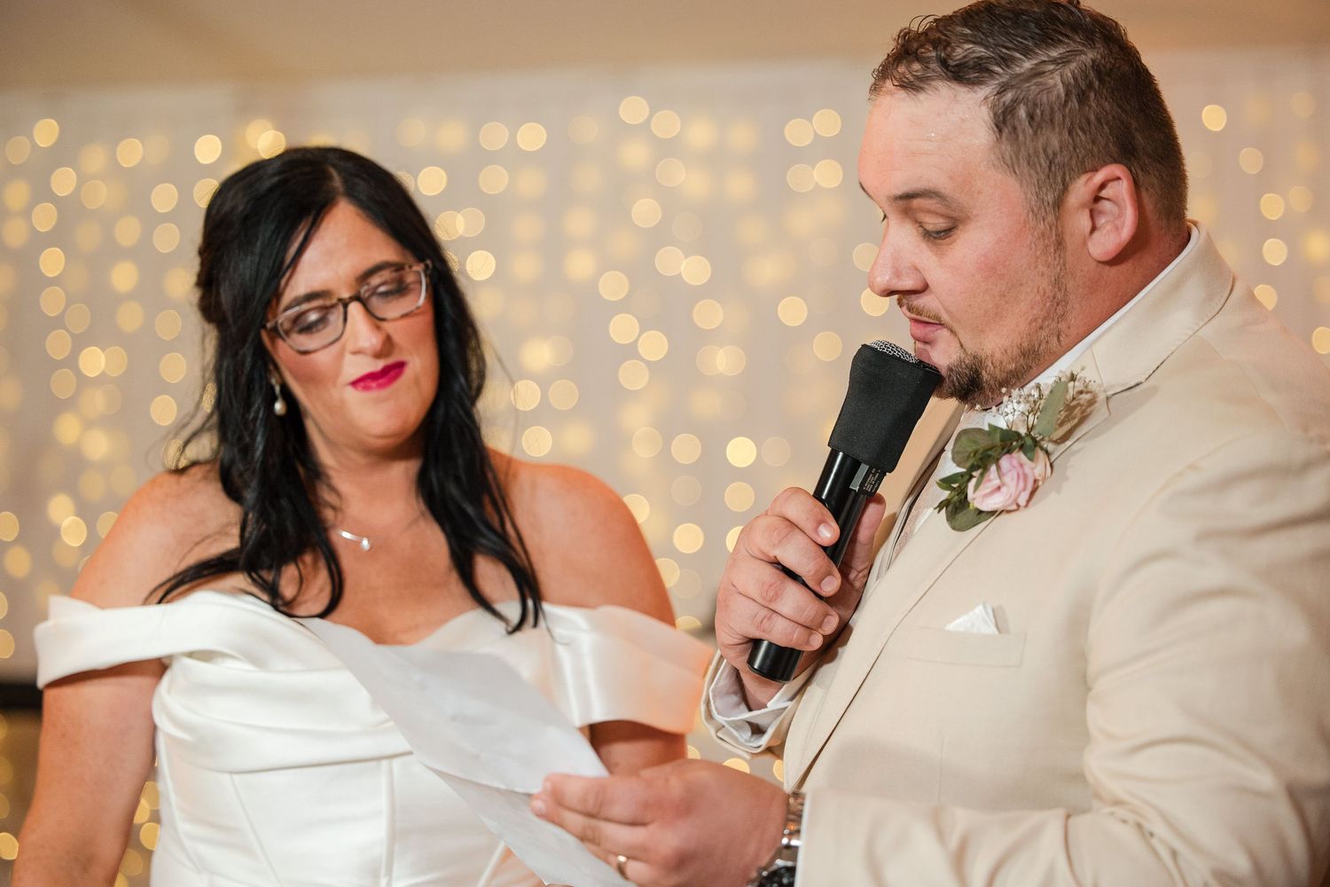 Bride and groom reading vows at wedding reception, groom holding microphone in white suit with floral boutonniere.