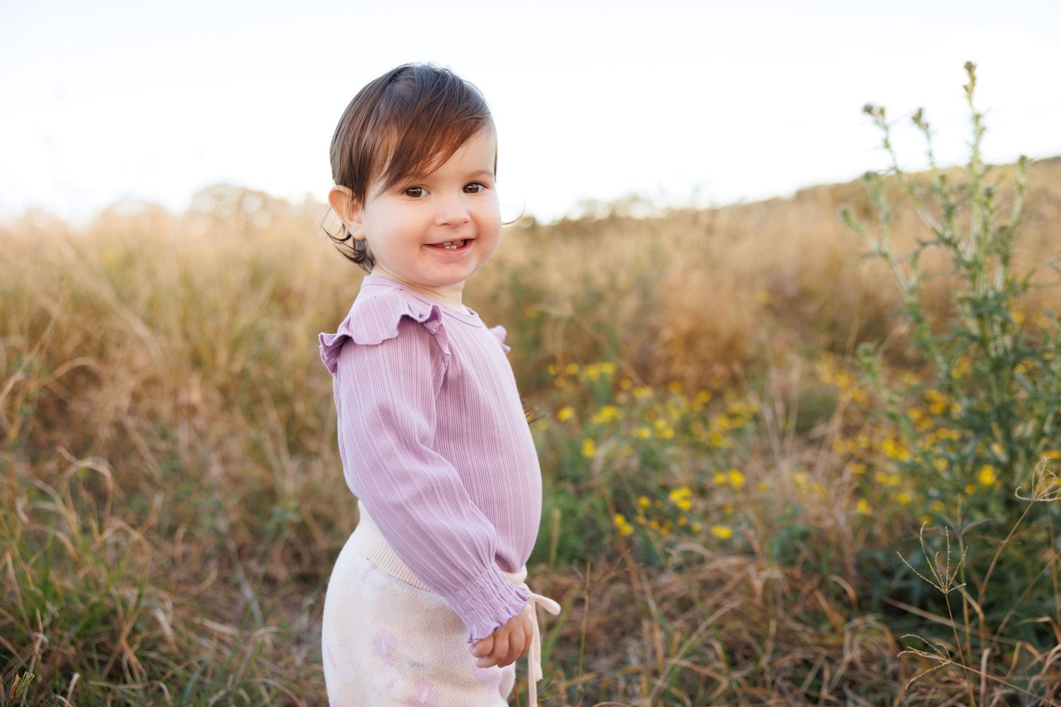 A toddler in a lavender shirt and white jumper dress smiles while playing in a golden field during sunset.