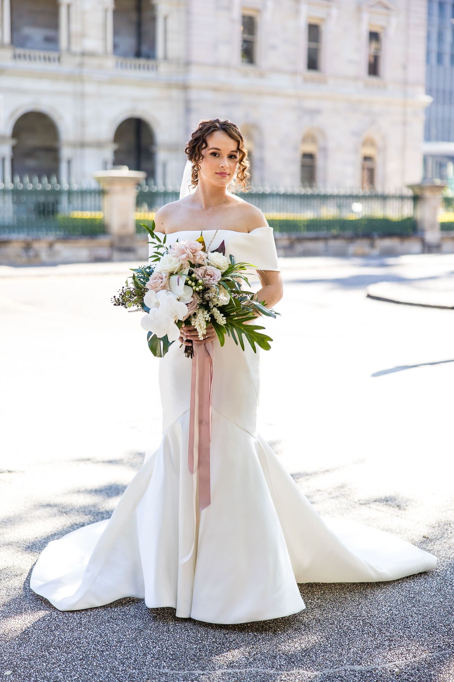 A bride in a classic white wedding gown holds a soft pink and white floral bouquet against an elegant architectural backdrop.