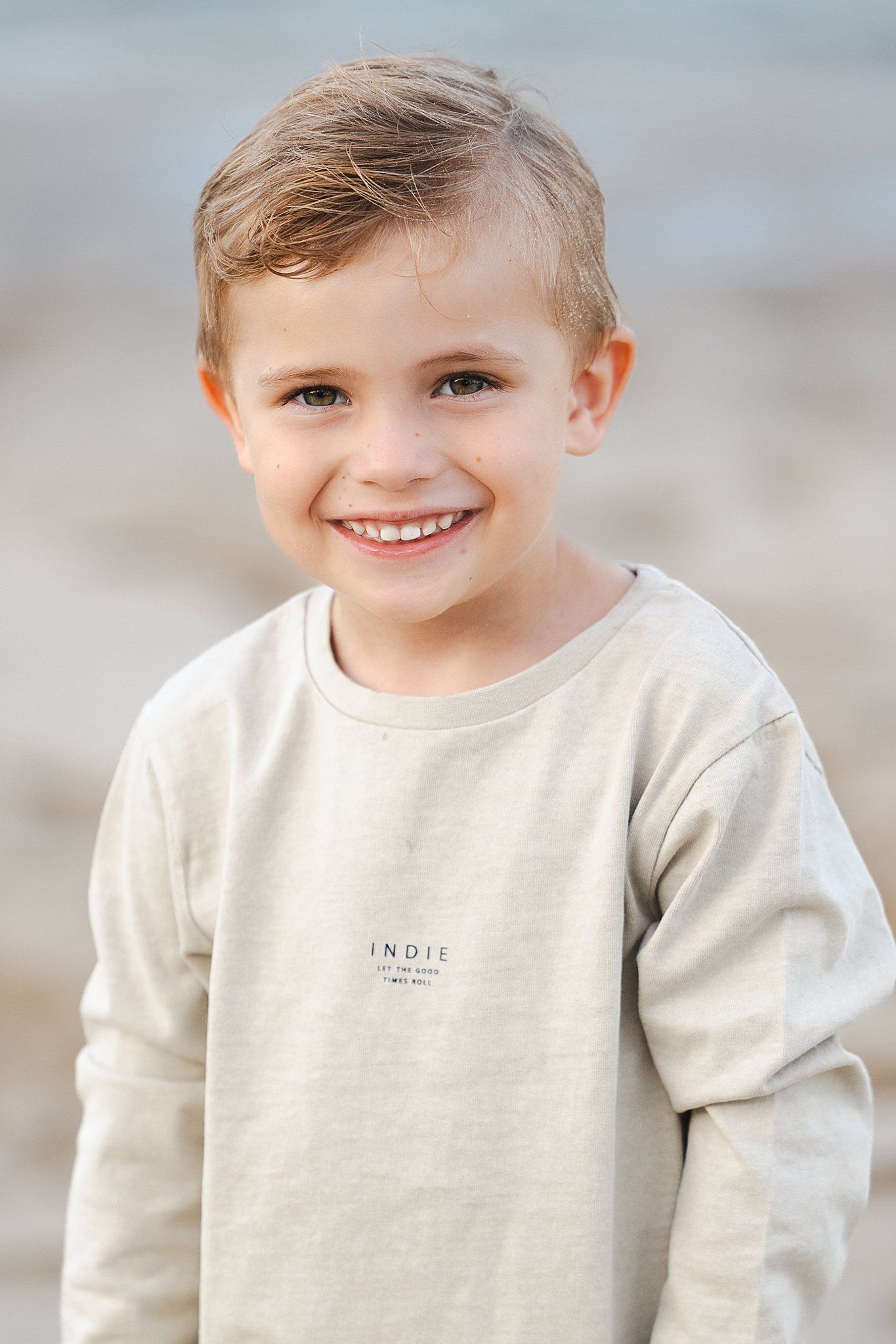 Child wearing a beige sweatshirt smiling at the camera against blurred background.