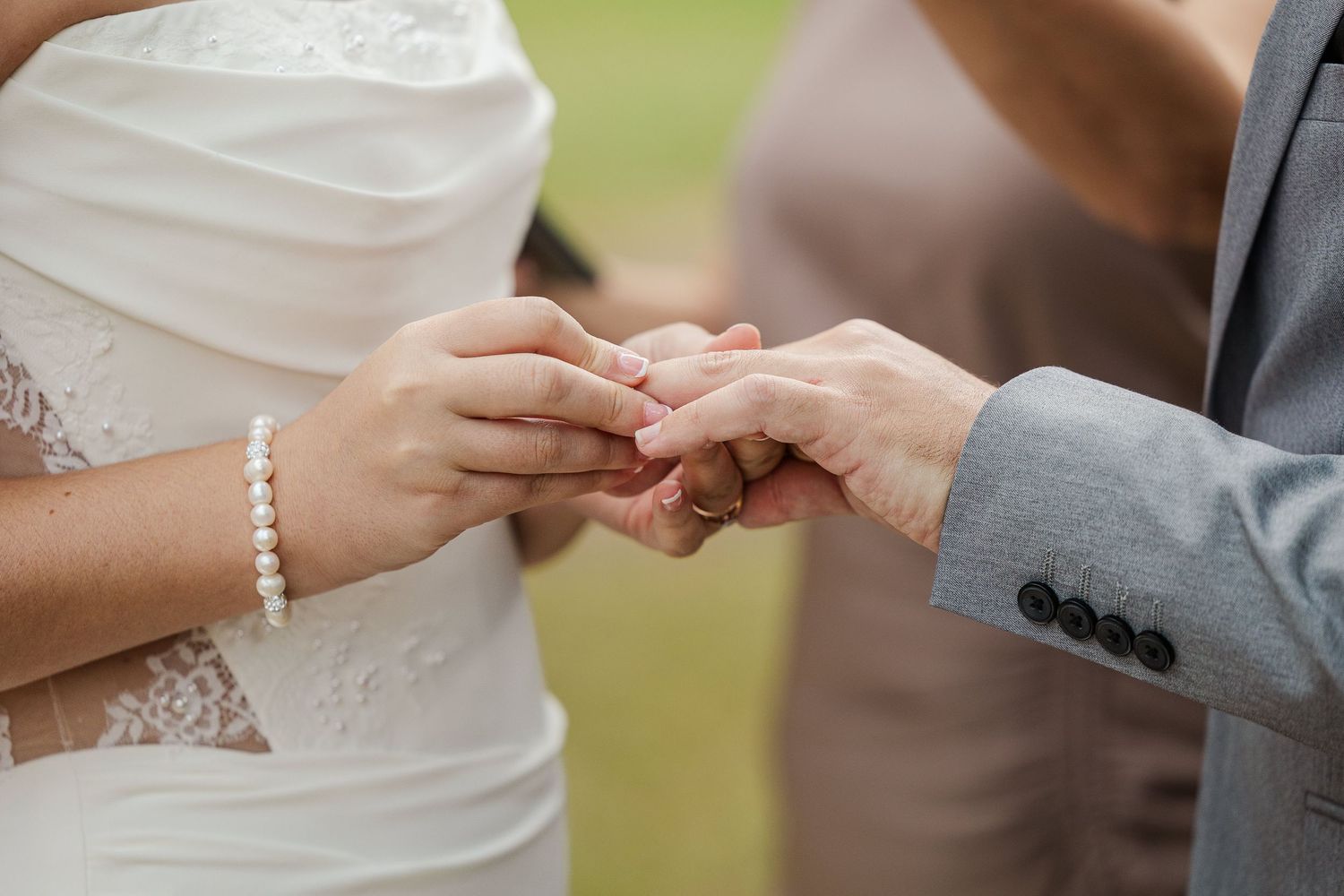 Close-up of bride and groom exchanging wedding rings during outdoor ceremony, bride wearing pearl bracelet.