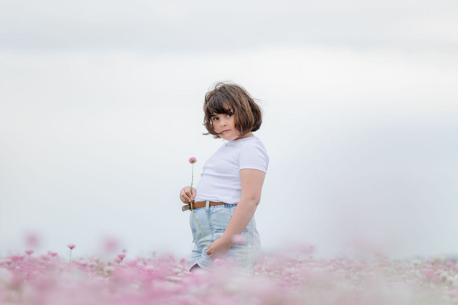 Young woman in white tee and jeans holding a flower in a pink cosmos field under a soft sky.