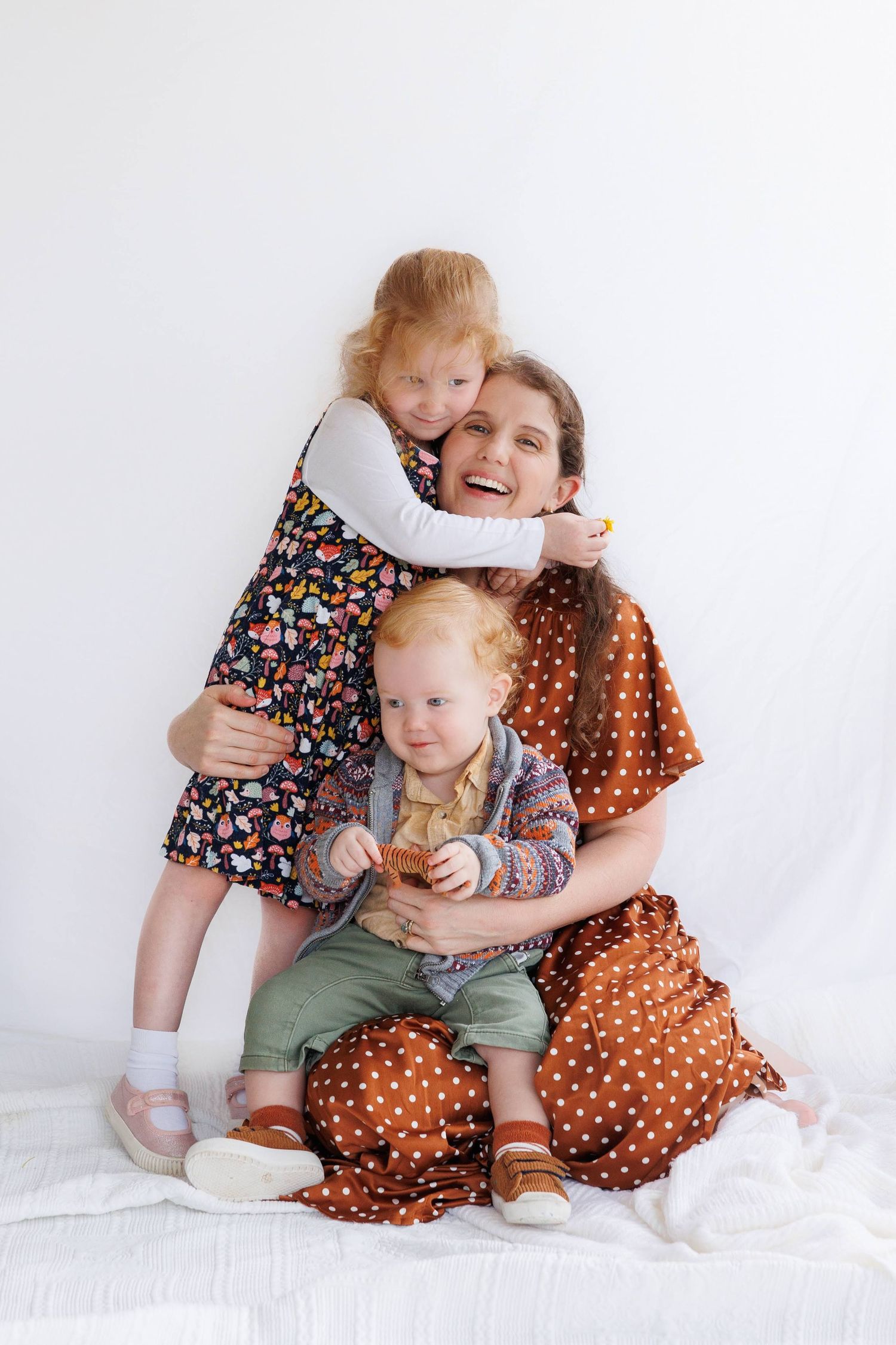 A joyful family portrait against a white backdrop features floral and polka dot dresses in warm earth tones.