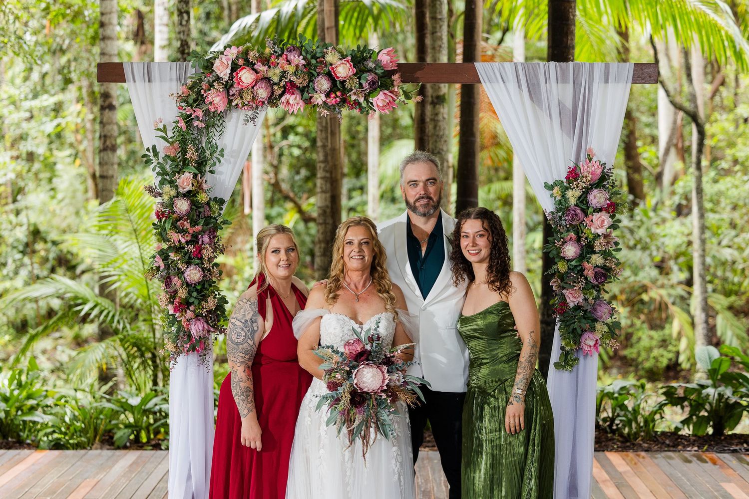 Bride and groom with guests posing under a floral arch with pink roses at an outdoor rainforest wedding ceremony.