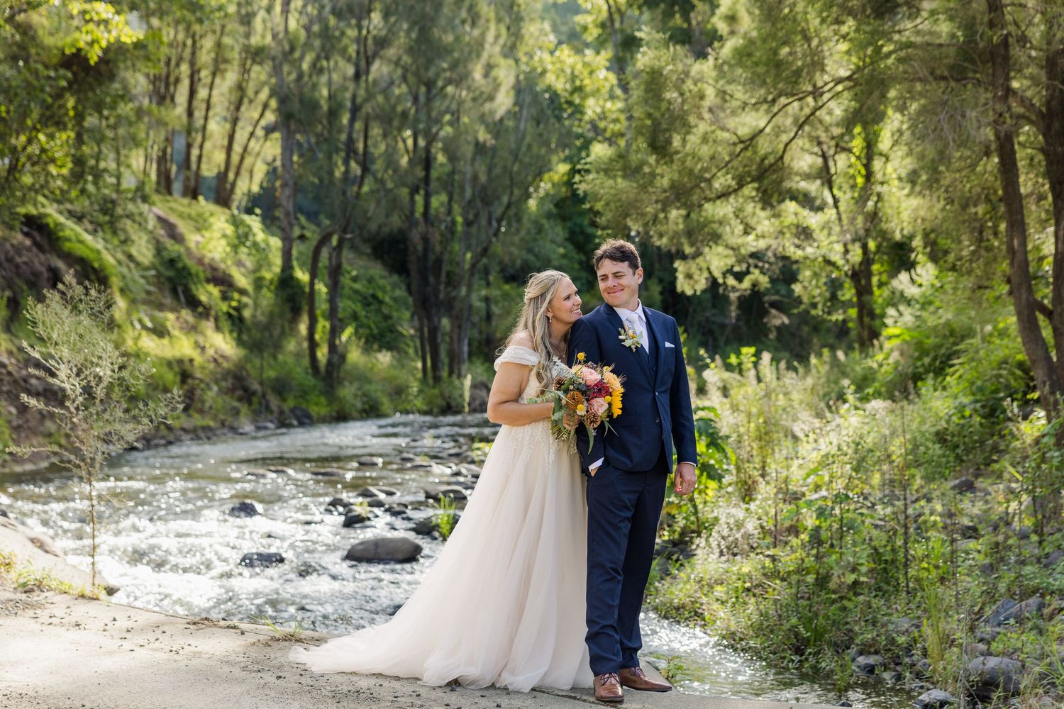 Bride and groom stand by a rocky creek surrounded by lush green trees, bride holding a colorful bouquet.