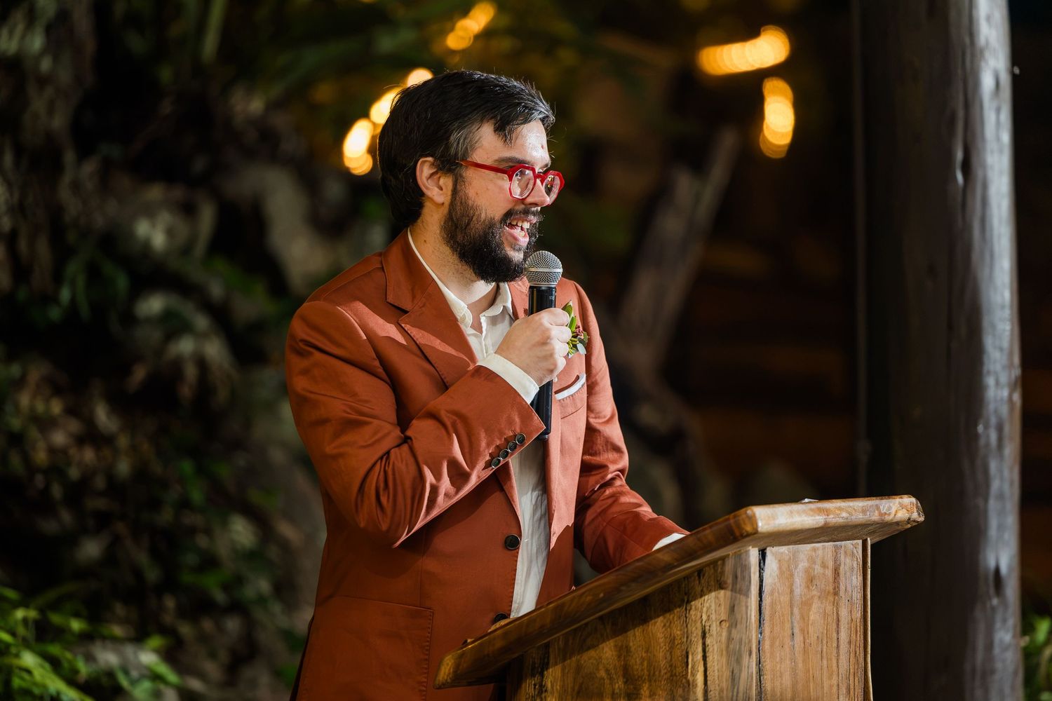 Man in red blazer and glasses speaking at podium with microphone at outdoor evening event.