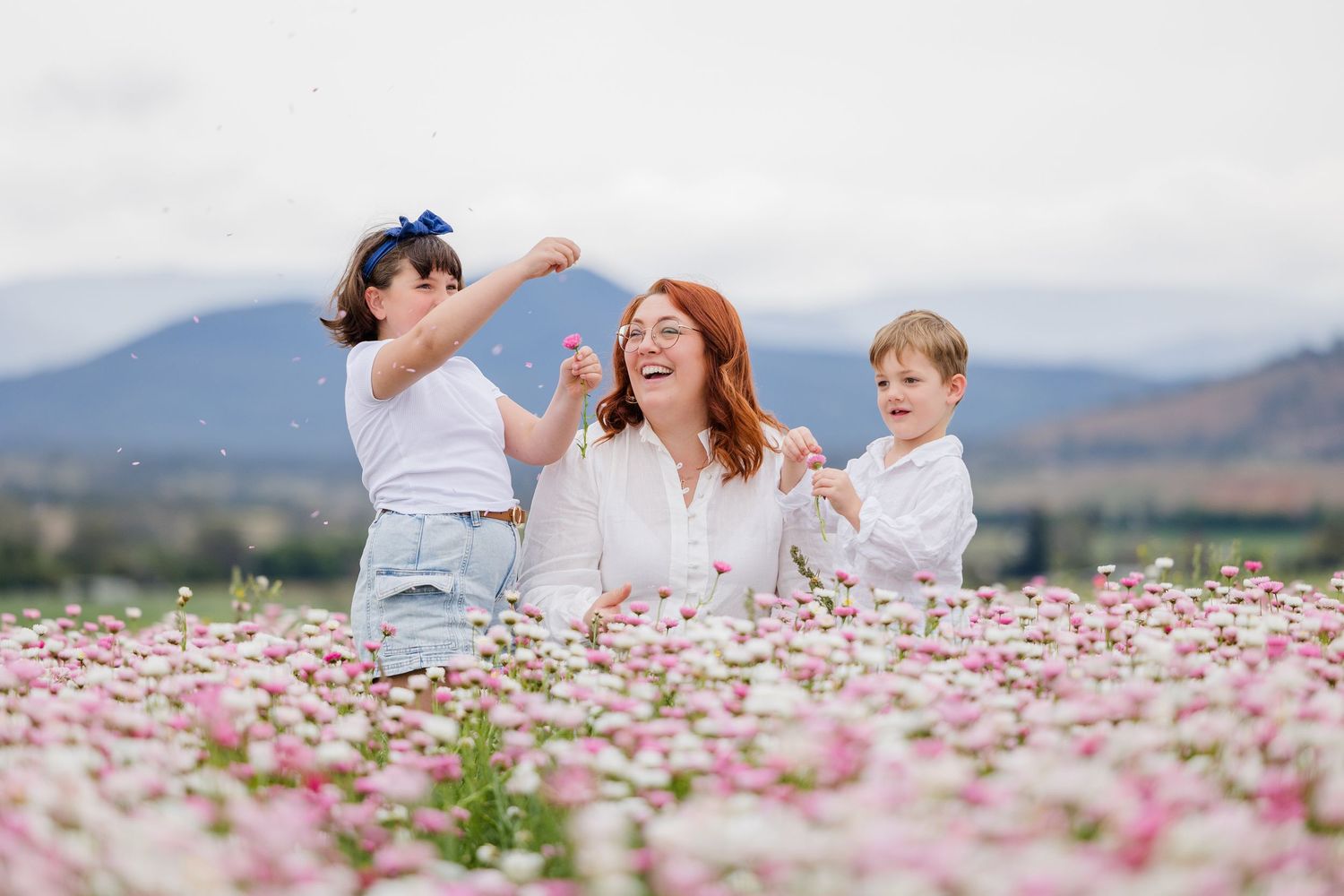 A joyful redhead mom laughs with two young children in a stunning pink and white flower field with mountains behind.