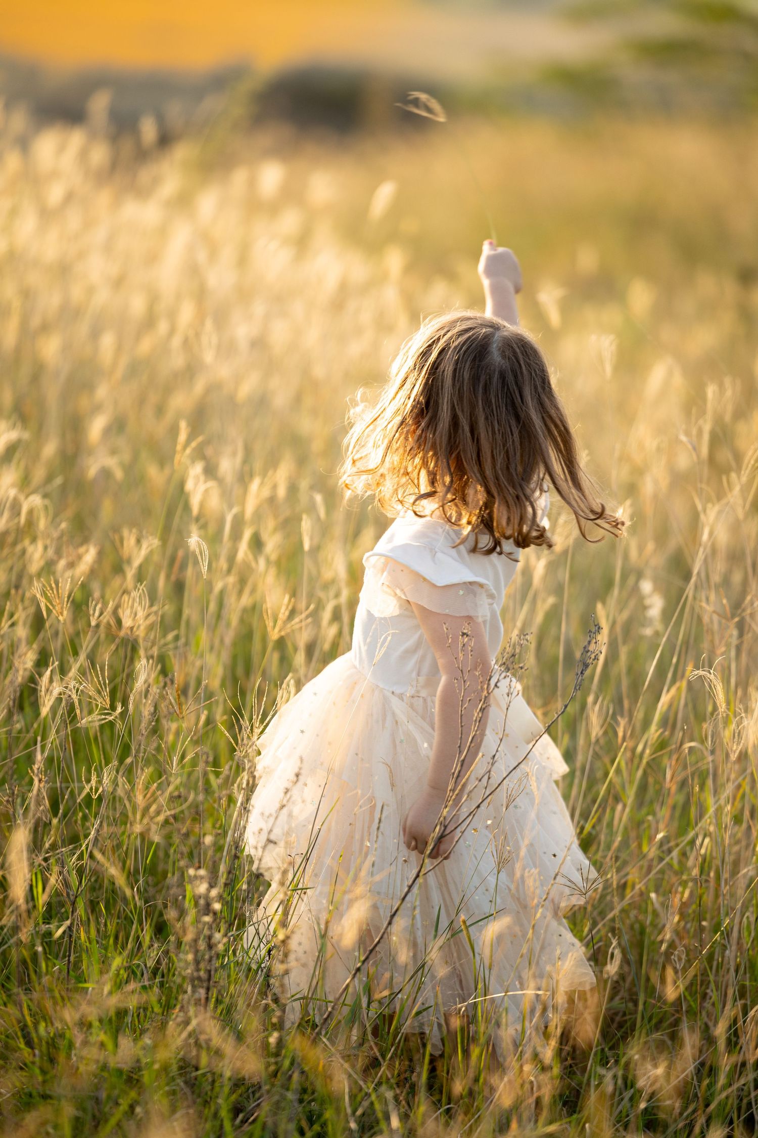 A young girl in a white dress twirls through a sun-lit field of tall golden grass during sunset.