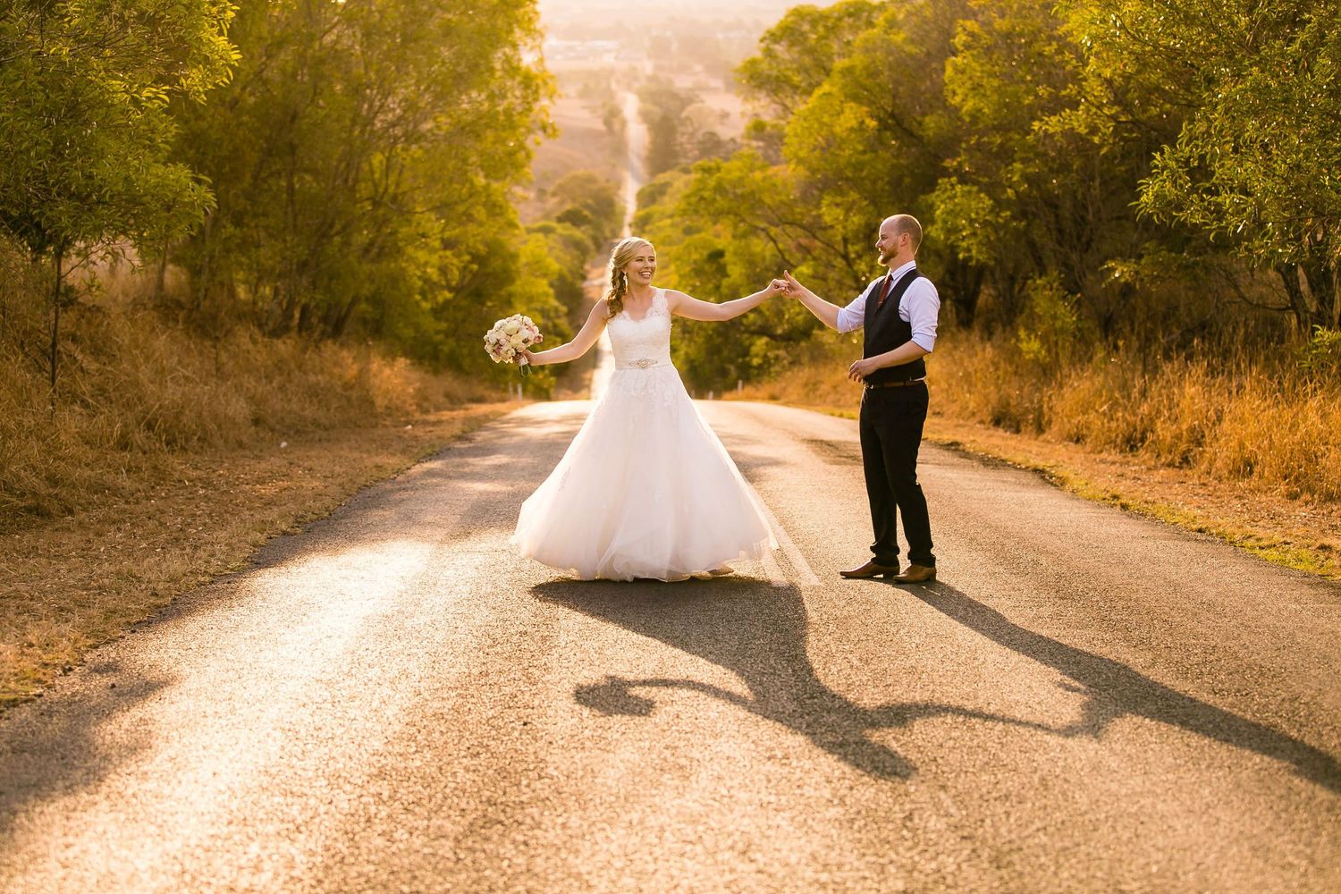 Newlyweds dancing on a sunlit country road surrounded by golden autumn trees.