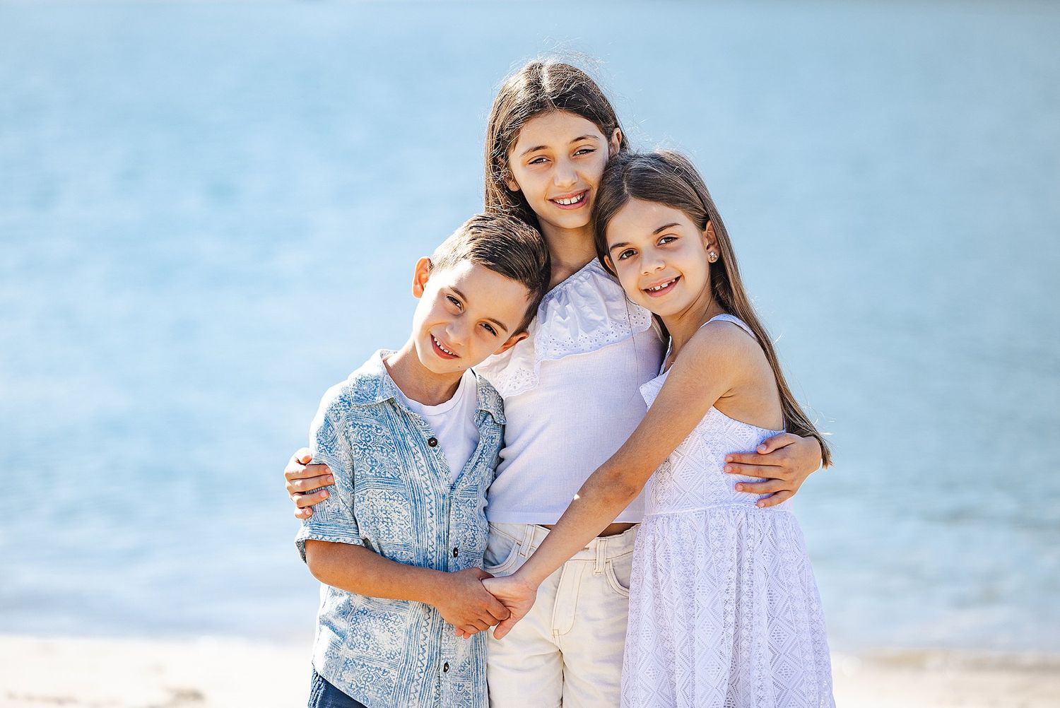 Three children wearing light summer outfits embrace while standing together on a sunny beach.