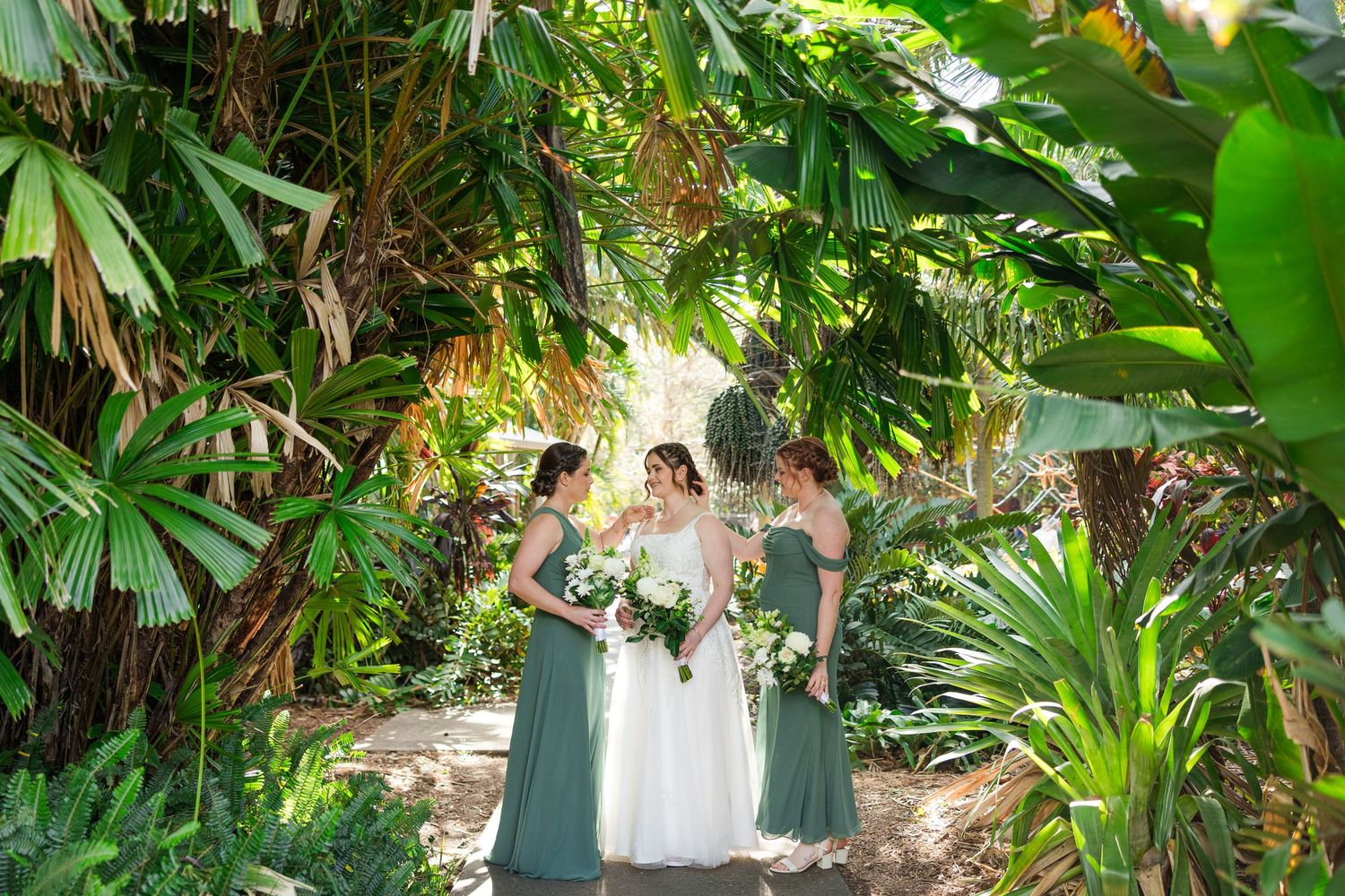 Bride and two bridesmaids in sage green gowns stand together among tropical palms and greenery.