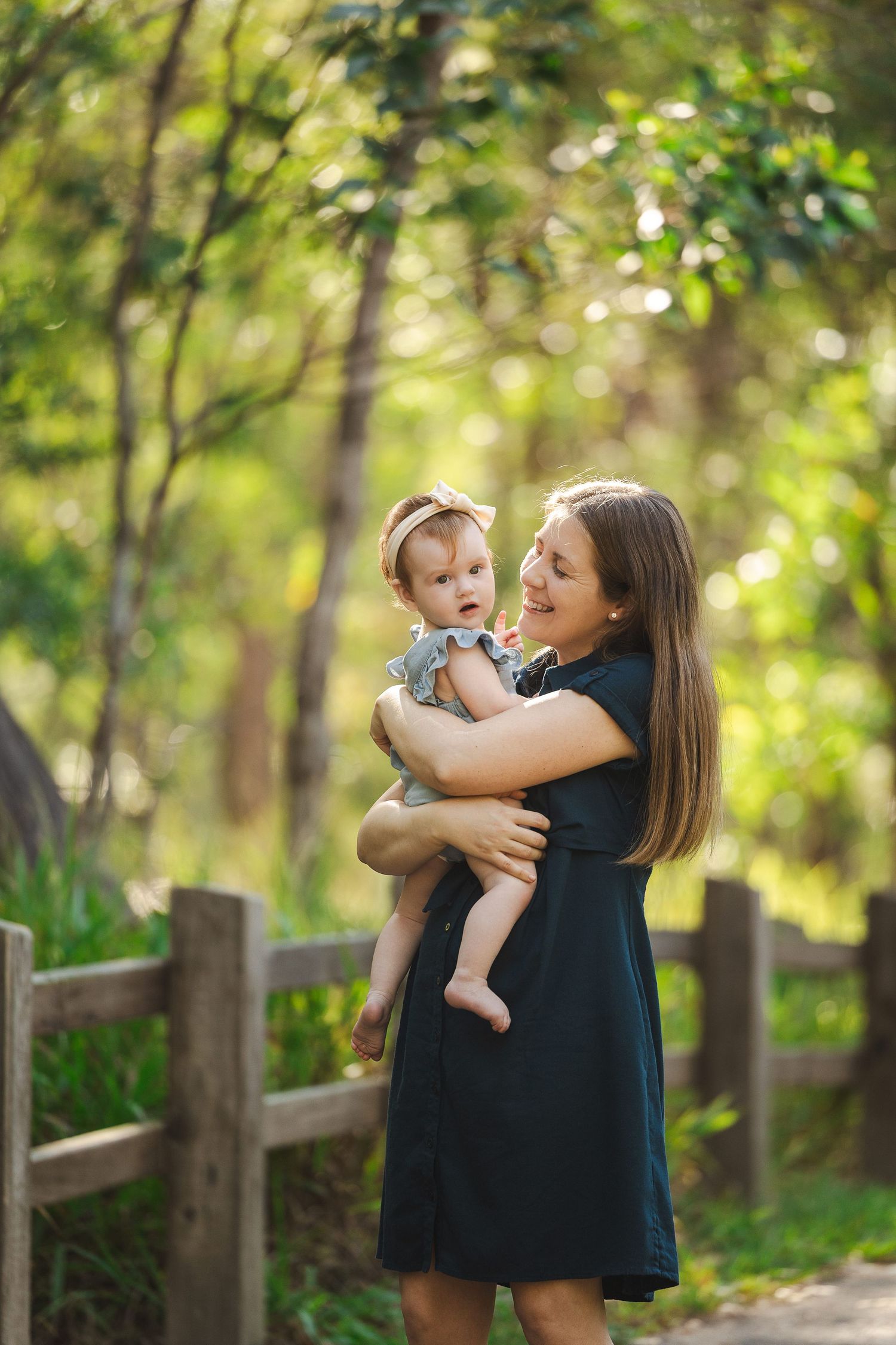 A tender moment shared between parent and baby in a sunlit wooded area with a rustic fence in the background.