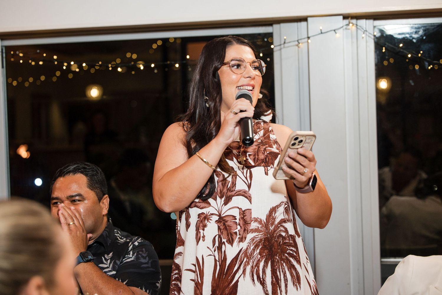 Woman in floral dress speaking into microphone at an indoor evening event with string lights.