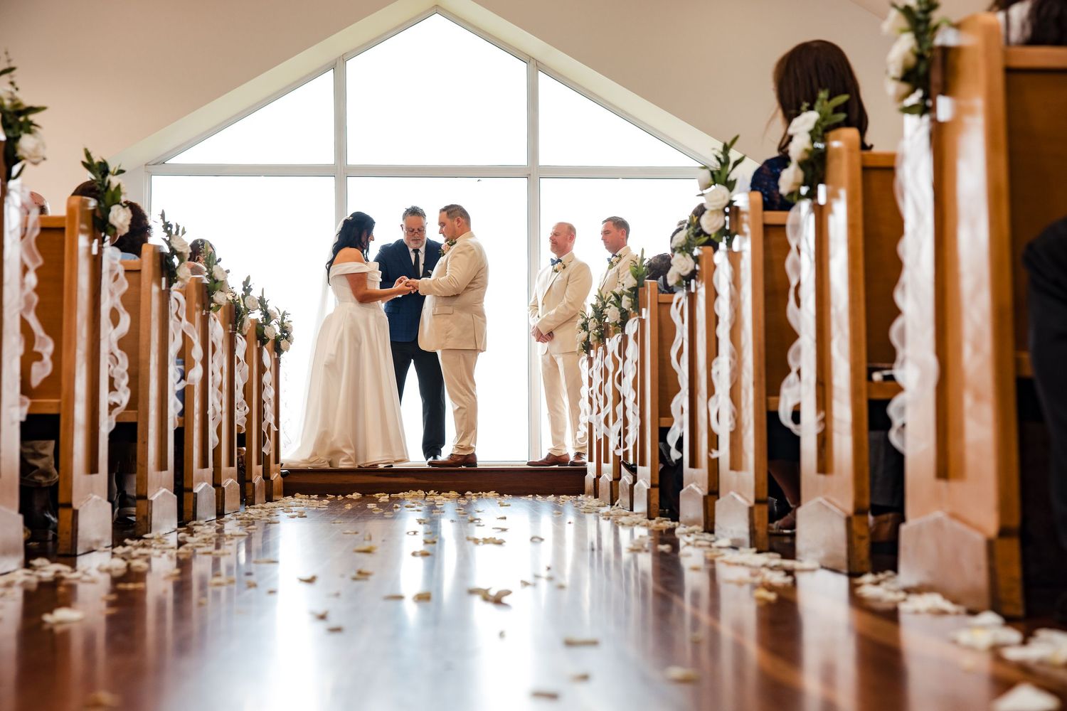 Elegant wedding ceremony in a chapel with floral-adorned pews, rose petals on aisle, couple exchanging vows at altar.