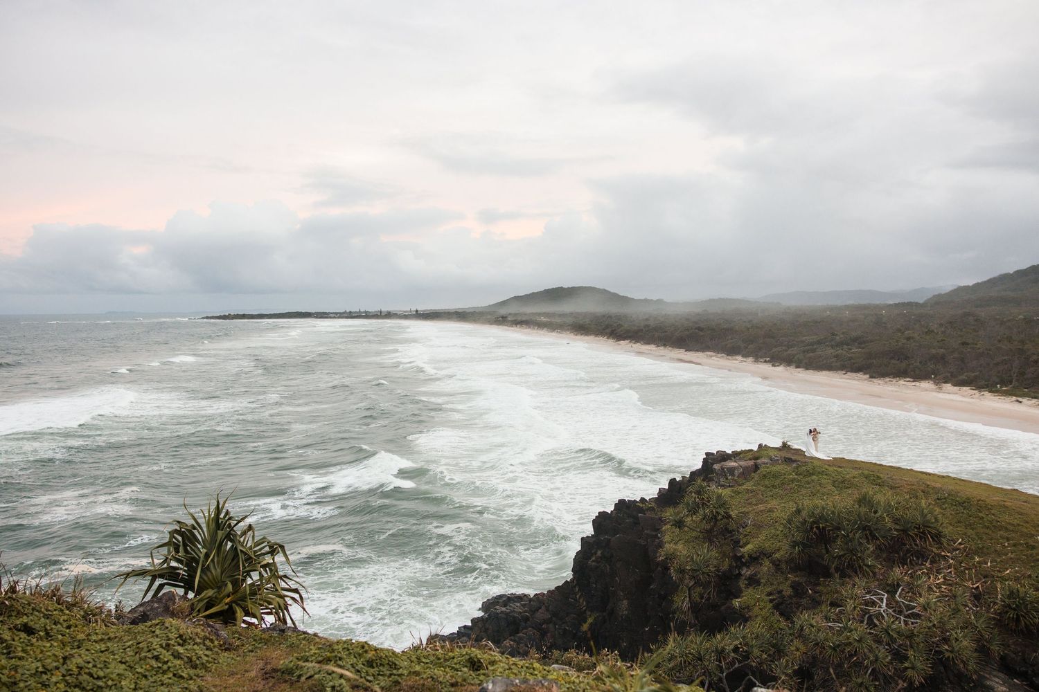 Aerial view of a rugged coastal beach with rolling waves, lush cliffs, and a hill under a cloudy sky.