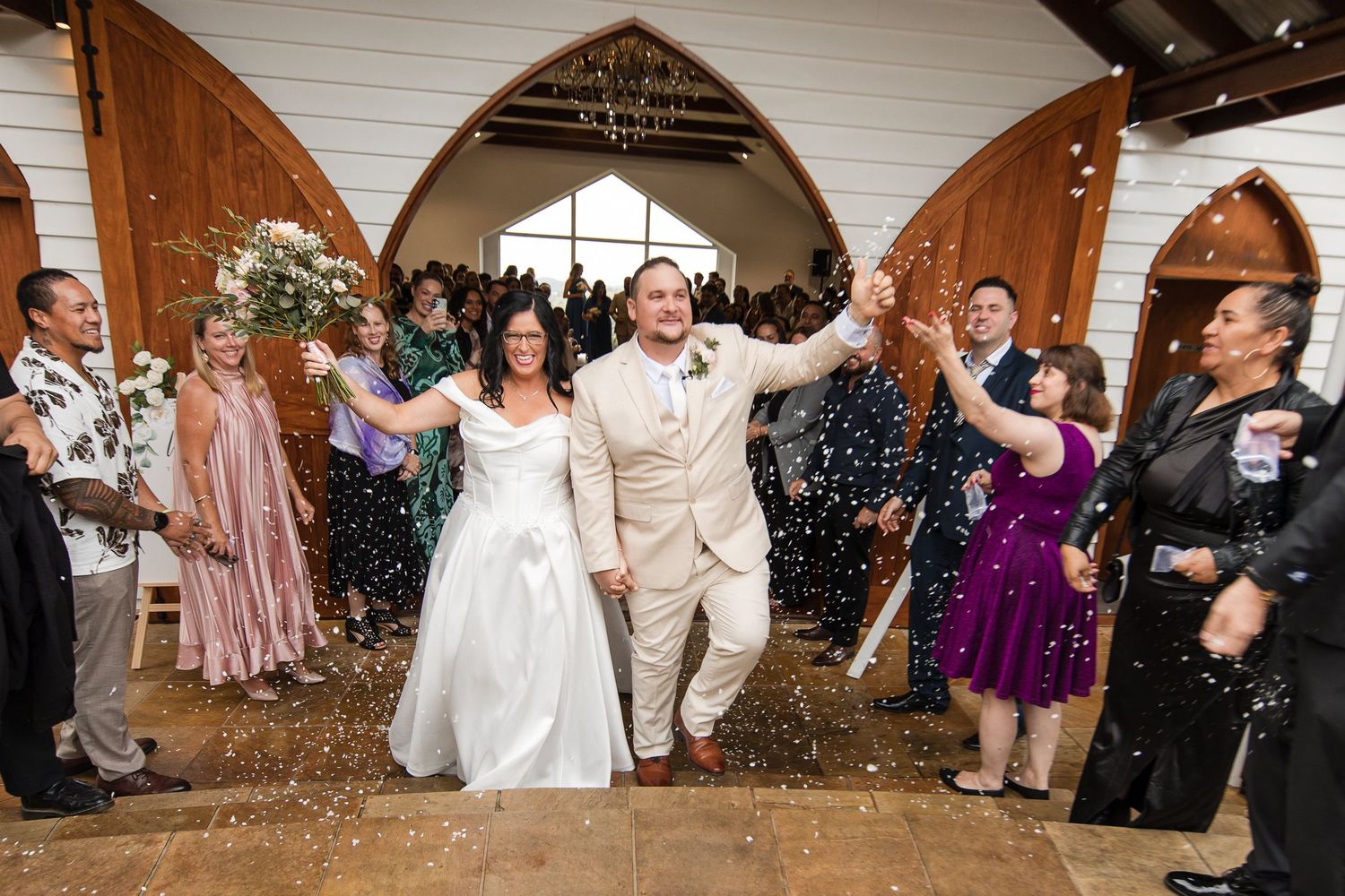 Bride and groom exit rustic chapel holding hands as guests toss confetti during joyful wedding celebration.
