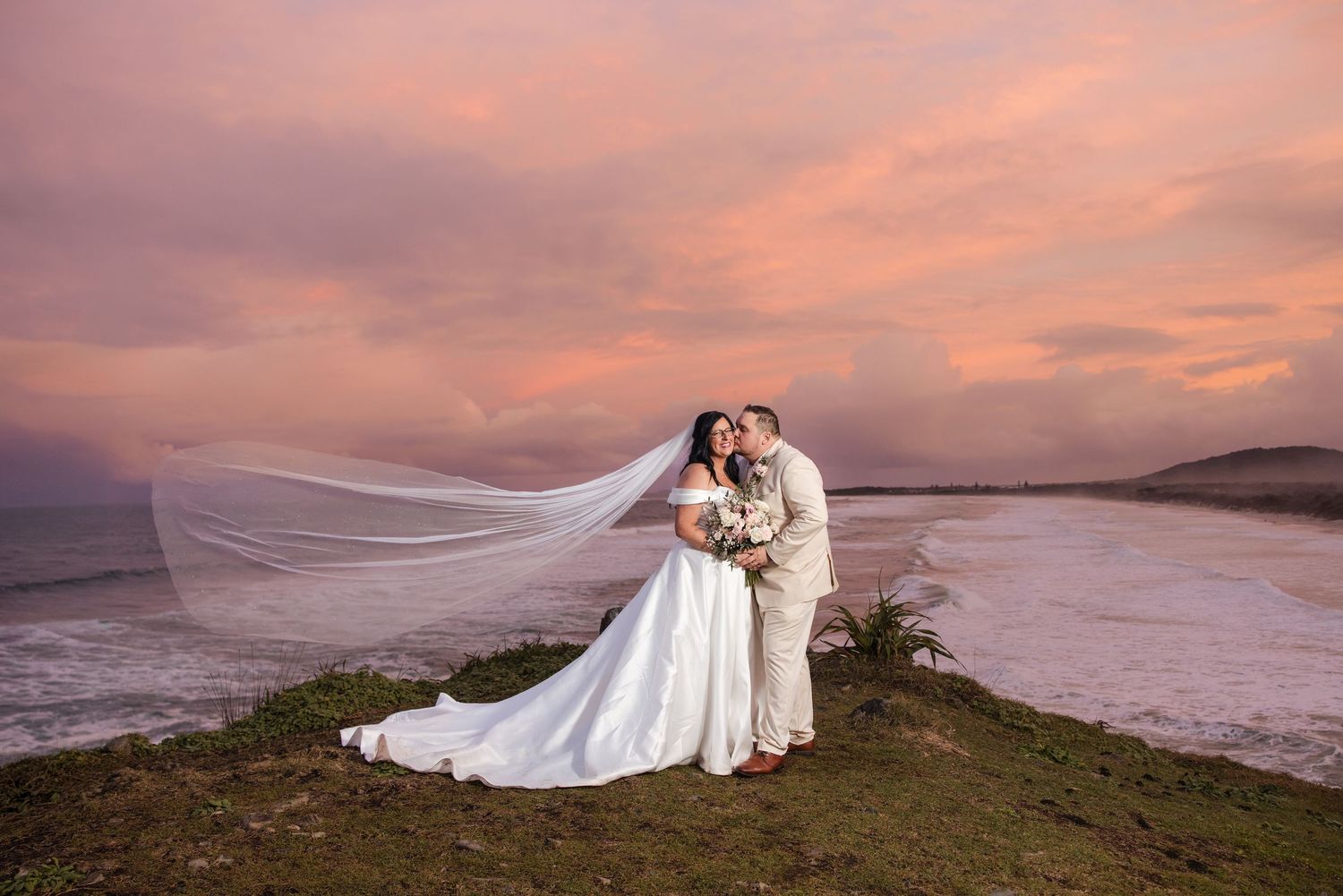 Bride and groom kissing on a coastal hill at sunset, with veil flowing in the wind against a pink sky.