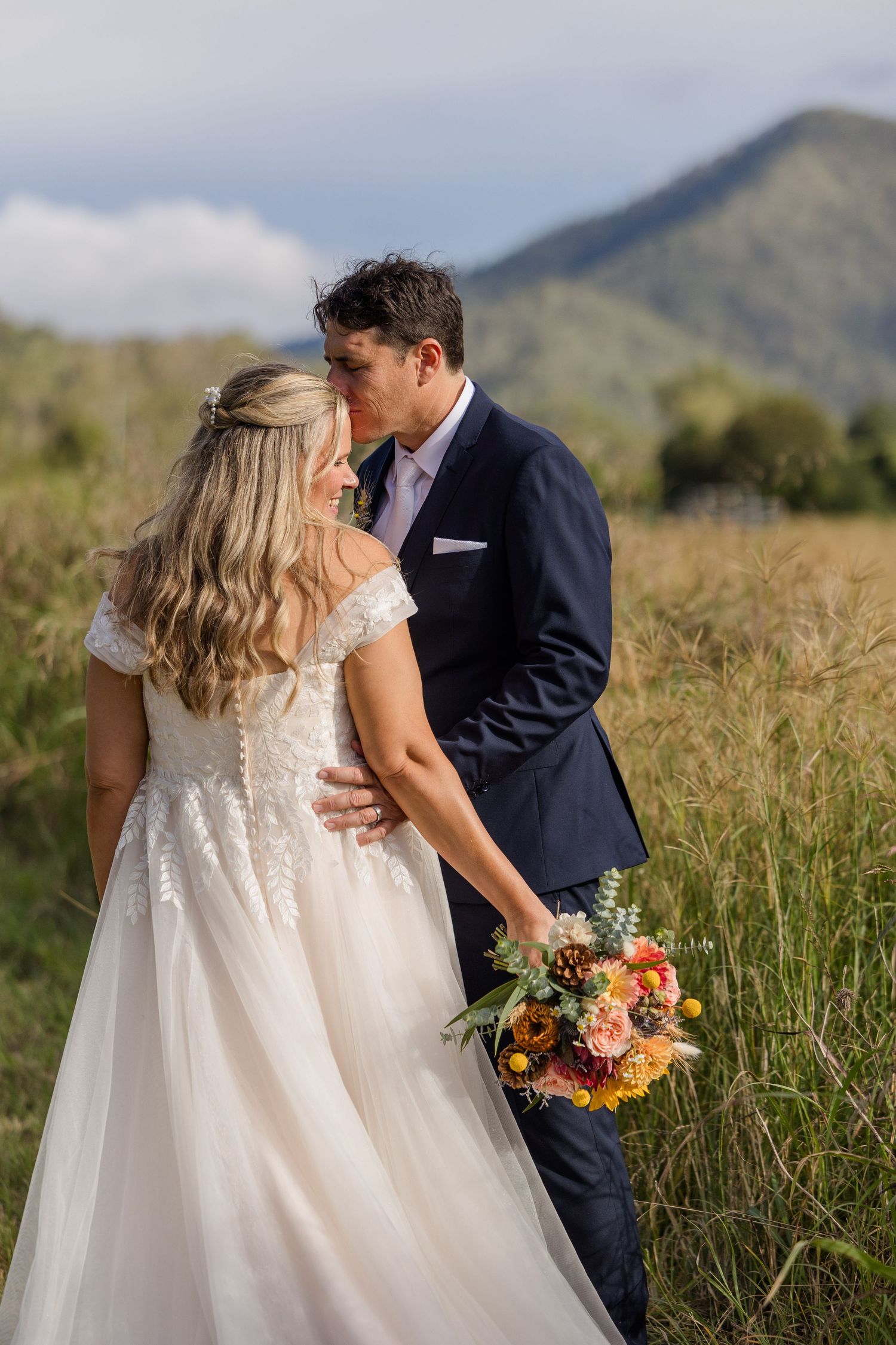 A bride and groom embrace in a field with mountains in the background, holding a bouquet of flowers.