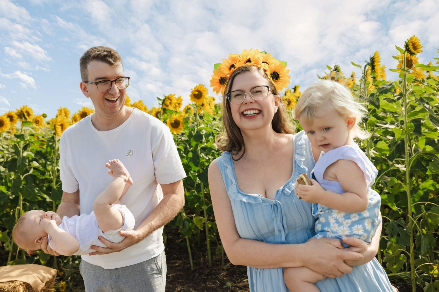 Family posing in sunflower field, woman wearing floral crown holding toddler, man holding baby, sunny day.