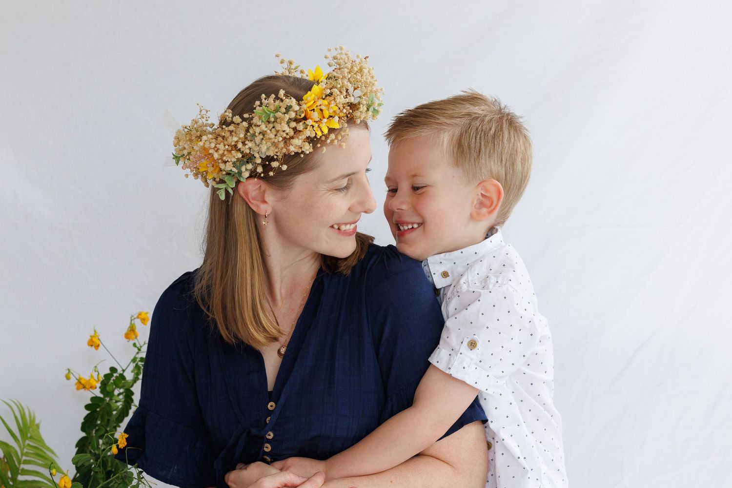 Mother wearing floral crown embraces young child in white shirt while sharing tender moment against light background.