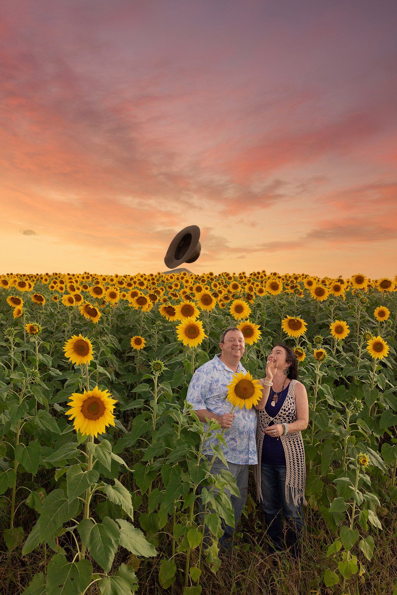 A couple poses in a vibrant sunflower field at sunset as a hat blows away in the golden sky.