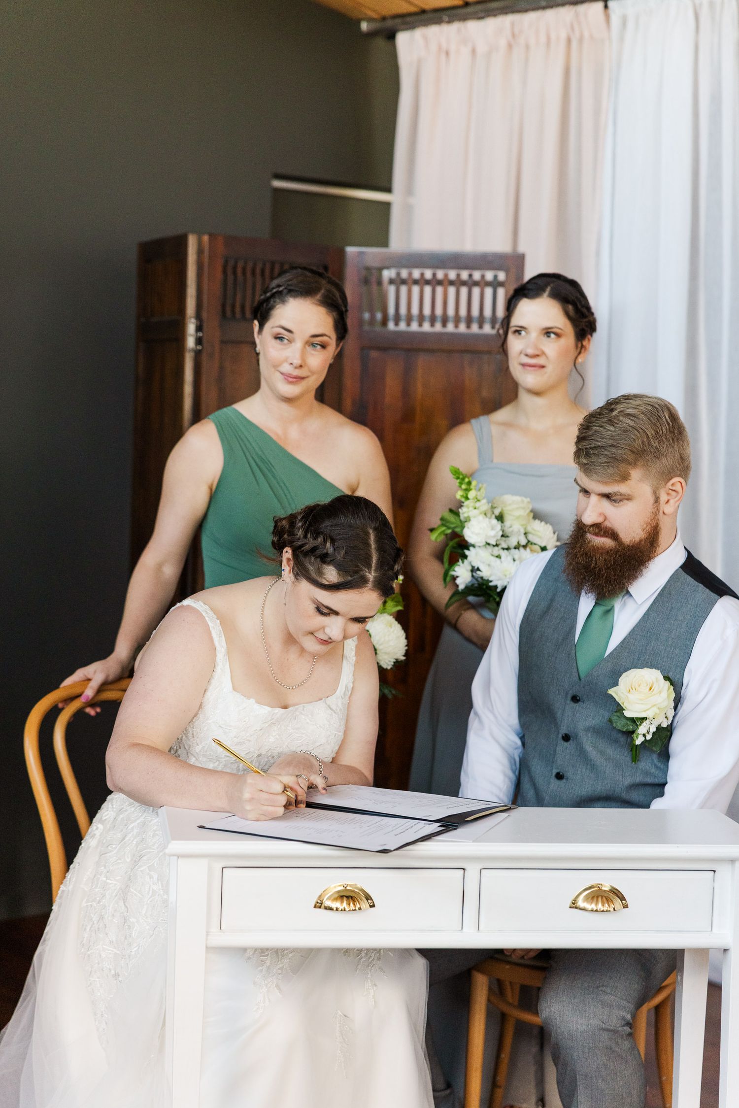 Bride signing wedding registry at white desk with groom and two bridesmaids in green and gray dresses.