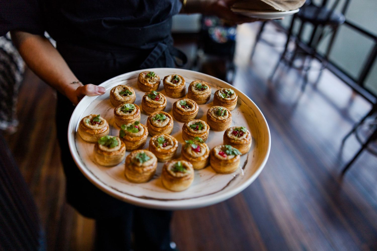 Server holding a round wooden platter of golden pastry appetizers topped with colorful garnishes at a catered event.