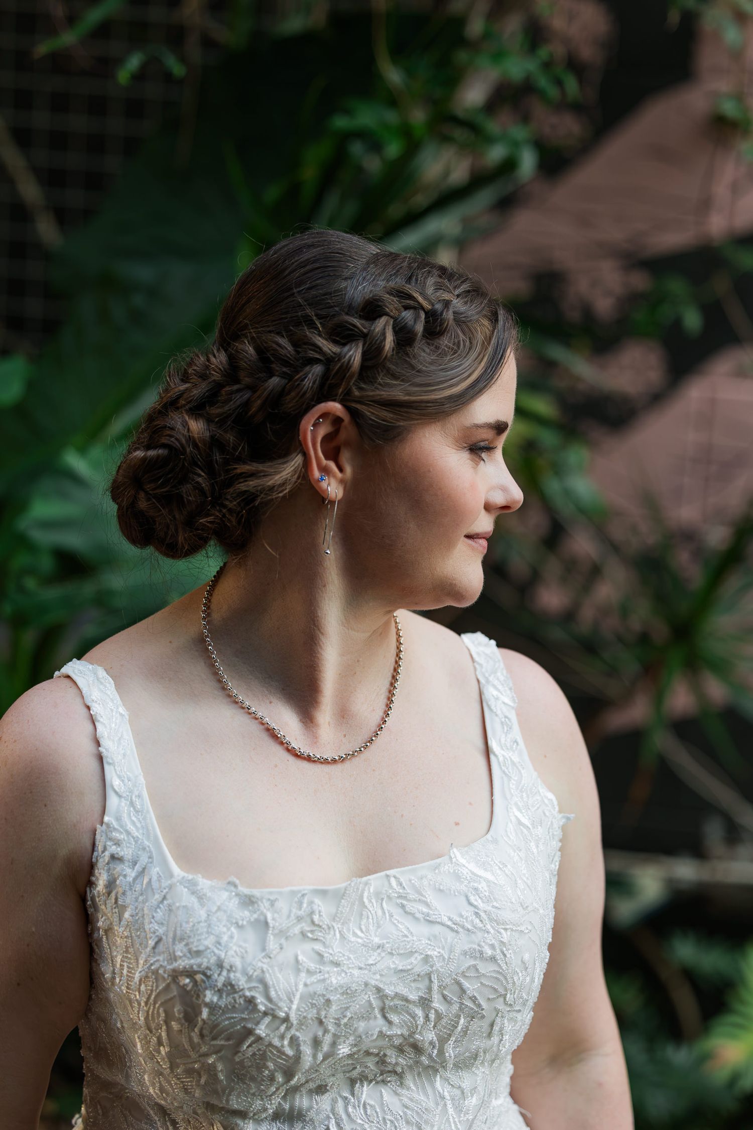 Bride in white lace dress with braided updo, pearl necklace, and earrings, posing outdoors with greenery background.