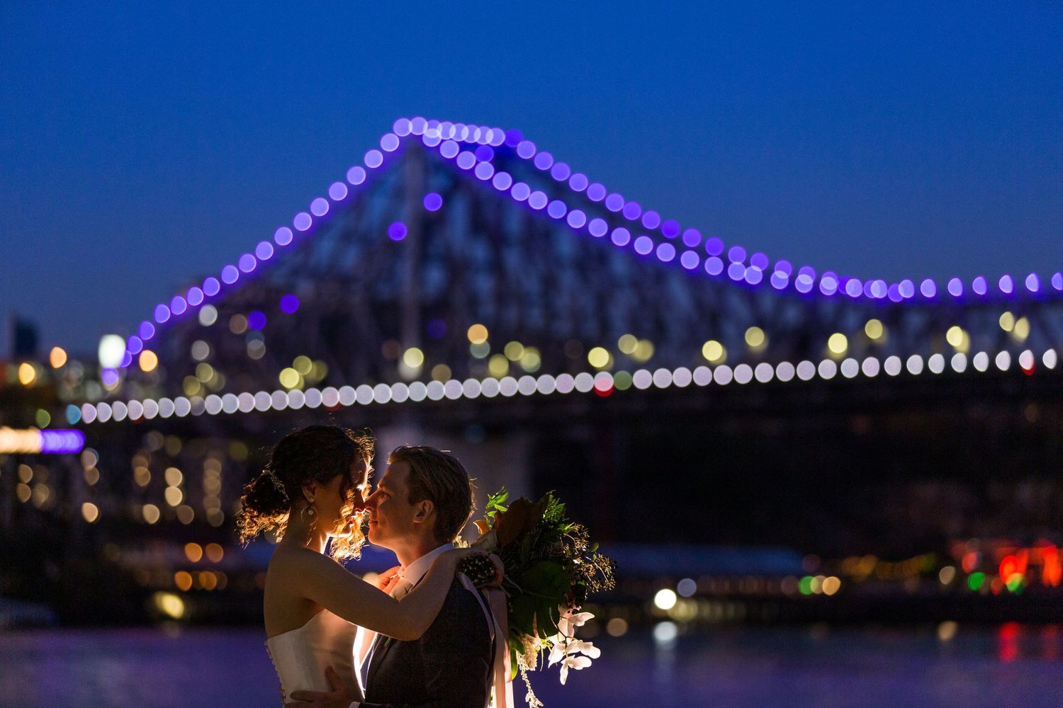 A romantic nighttime wedding photo with the Story Bridge lit up in purple in Brisbane.