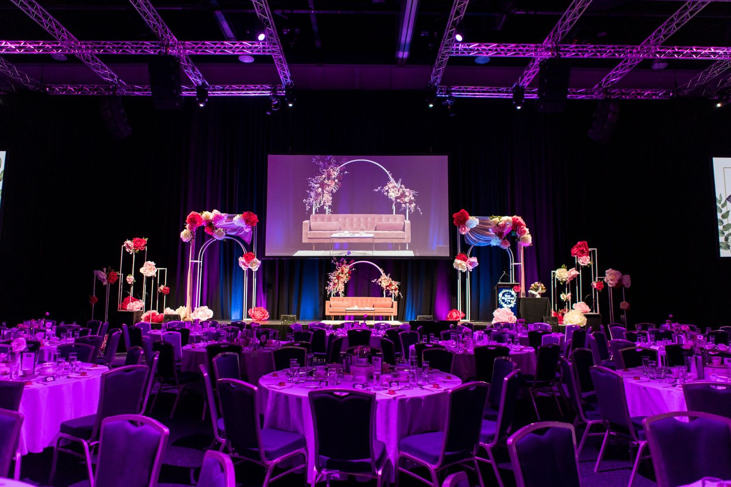 Elegant banquet hall setup with purple lighting, white chairs, floral centerpieces and a decorative stage display.